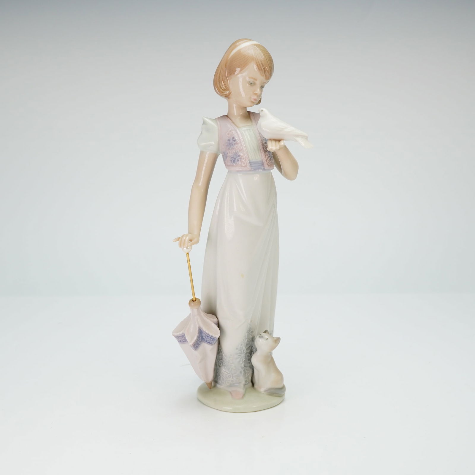 Lladro Porcelain Figurine, Summer Stroll 01007611: A Lladro glazed porcelain figurine depicting a young woman in a long white gown with floral hem detailing and a pink patterned vest. The figure is shown holding a white dove and a parasol, with a kitt