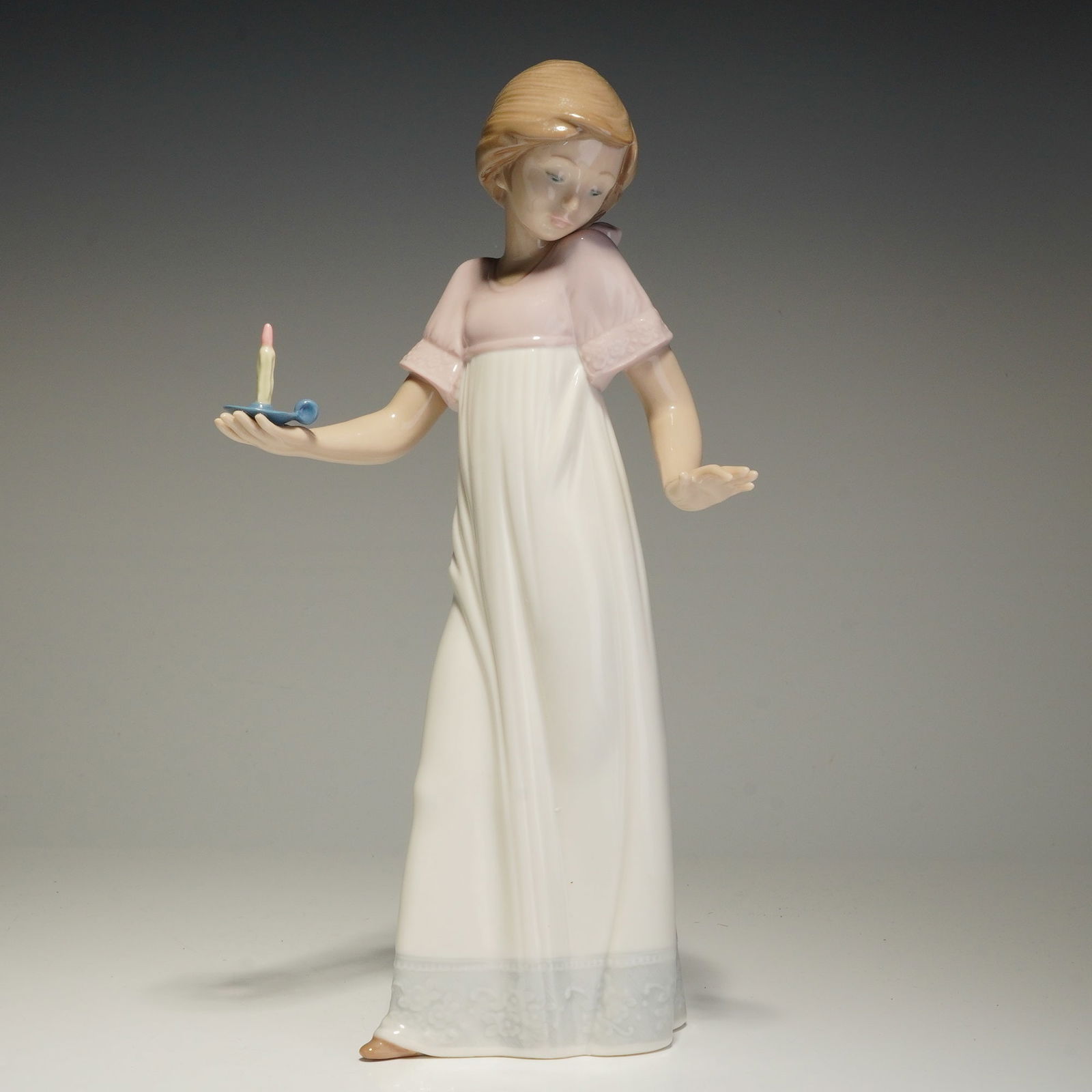 Nao by Lladro Porcelain Figurine, To Light The Way: The figure depicts a young girl standing in a full-length white nightgown featuring a pink yoke and a matching pink bow in her light brown hair. She is posed holding a blue candlestick with a yellow c