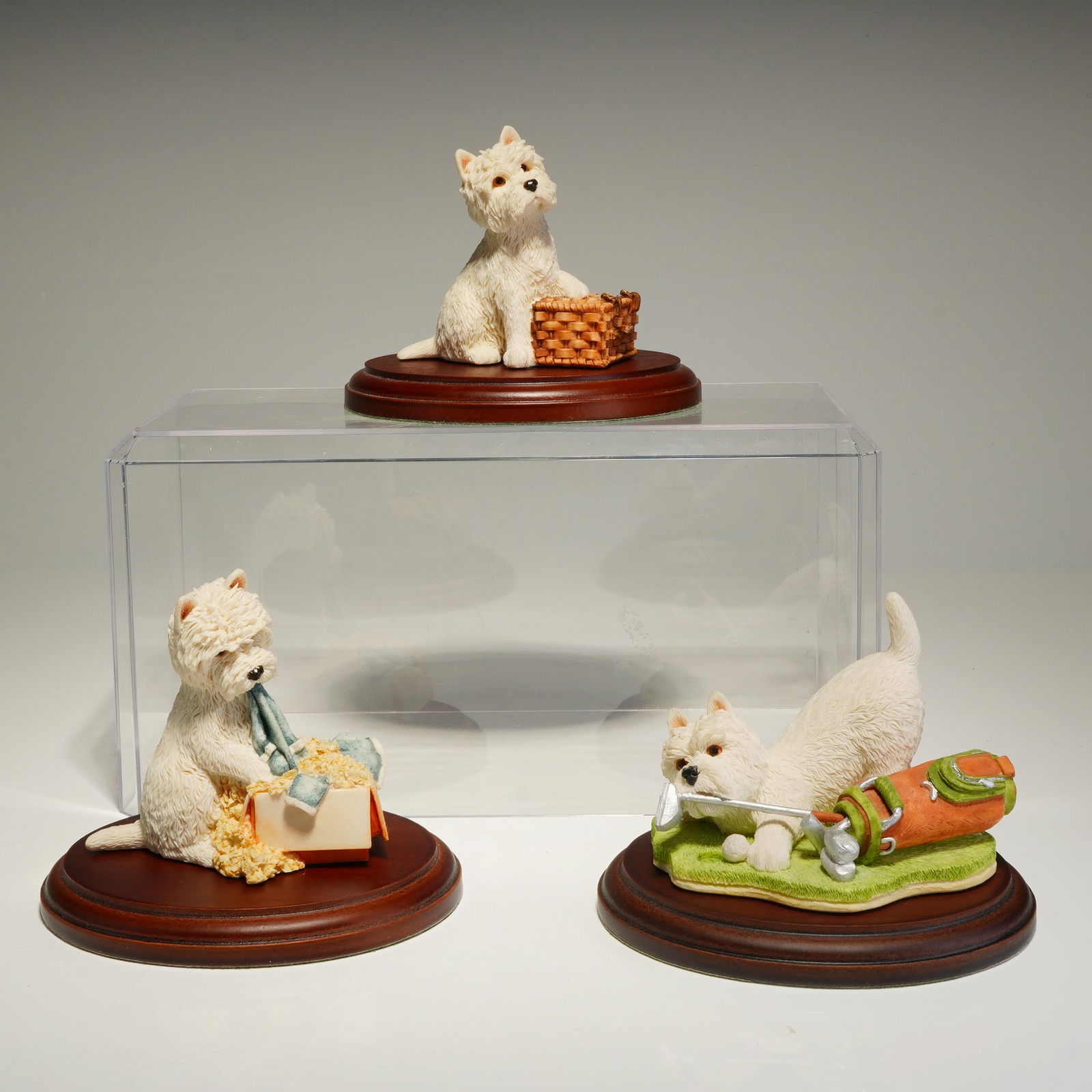 Trio of Sherratt & Simpson West Highland White Terrier Figurines with Bases: The group consists of three distinct poses: one terrier with a woven basket, one with a laundry box containing garments, and one on a green-colored base with a golf bag and club. The figurines are