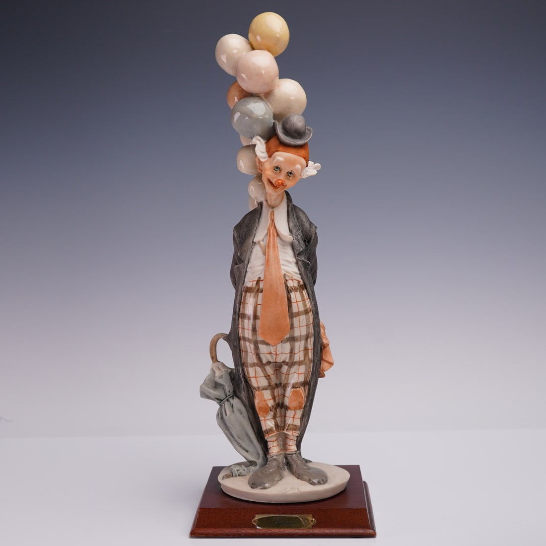Pucci Clown Figurine with Balloons on Wood Base: Tall porcelain clown figurine by Pucci, featuring a lanky character in plaid pants and oversized tie holding a whimsical bouquet of pastel balloons above his head. The figure is mounted to a polished