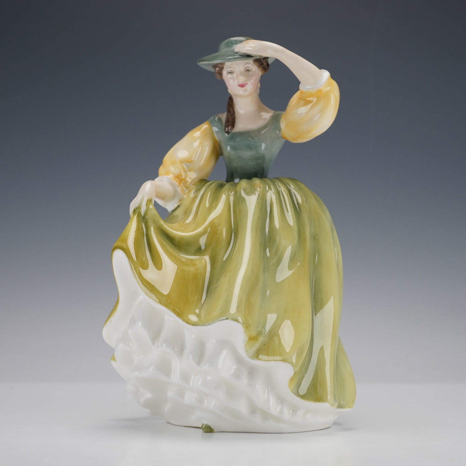 Royal Doulton Figurine, Buttercup HN2309: A glazed porcelain modeled in green gown with yellow golden sleeves. She holds her matching hat from blowing away. Royal Doulton backstamp. Artist: Peggy Davies Issued: 1964 - 1997 Dimensions: 4.75"L
