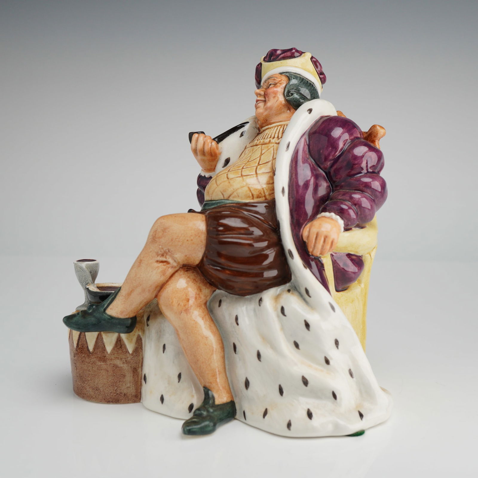 Royal Doulton Figurine, Old King Cole HN2217: A Royal Doulton ceramic figurine titled Old King Cole, model HN 2217. The glazed porcelain figure is depicted in a seated position within a high-backed yellow chair, wearing a purple and white ermine-