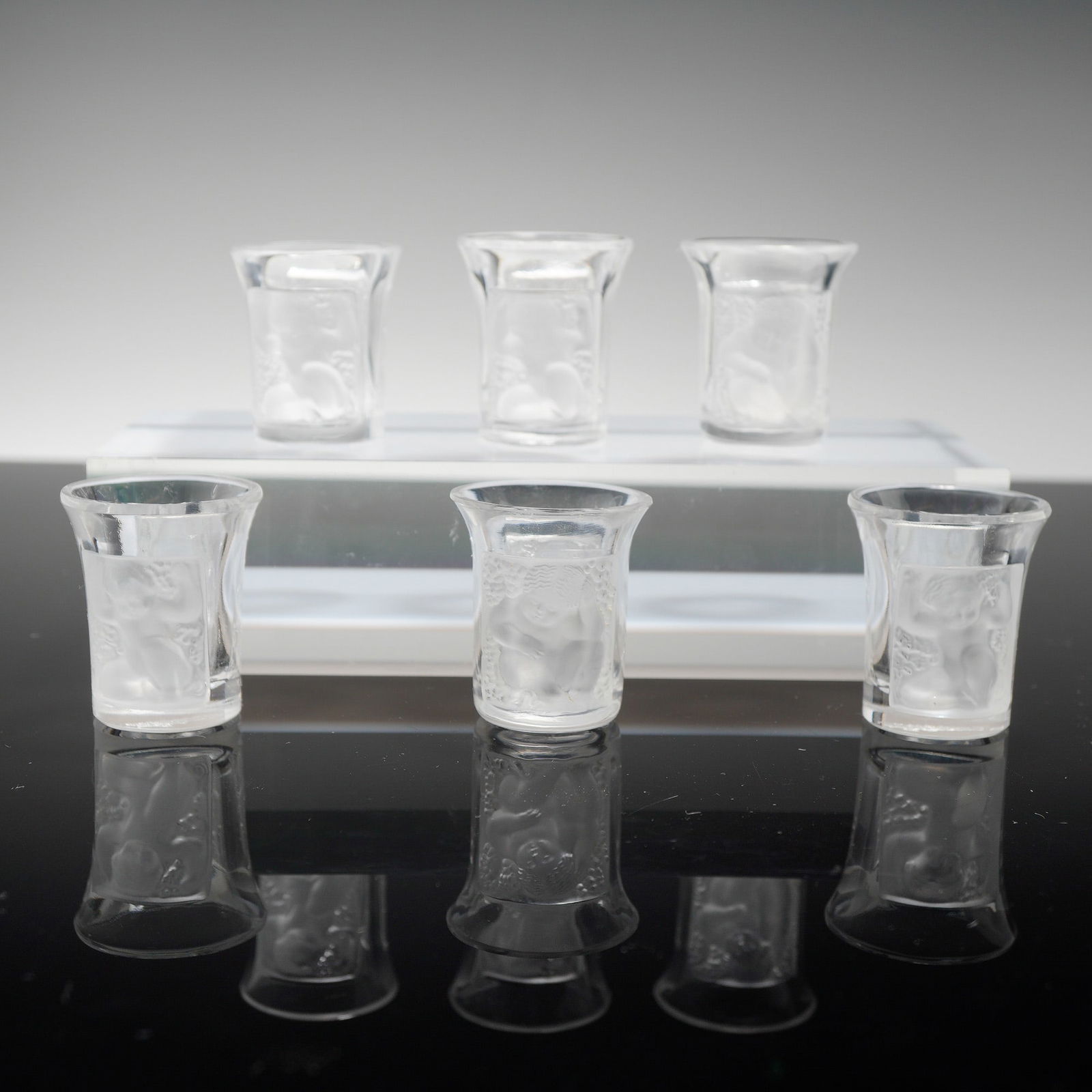 Set of Six Lalique France Crystal Les Enfants Shot Glasses: Each vessel is constructed from clear crystal featuring frosted rectangular panels with relief-molded designs of cherubs or children amongst foliage. The glasses are designed with a slightly flared ri