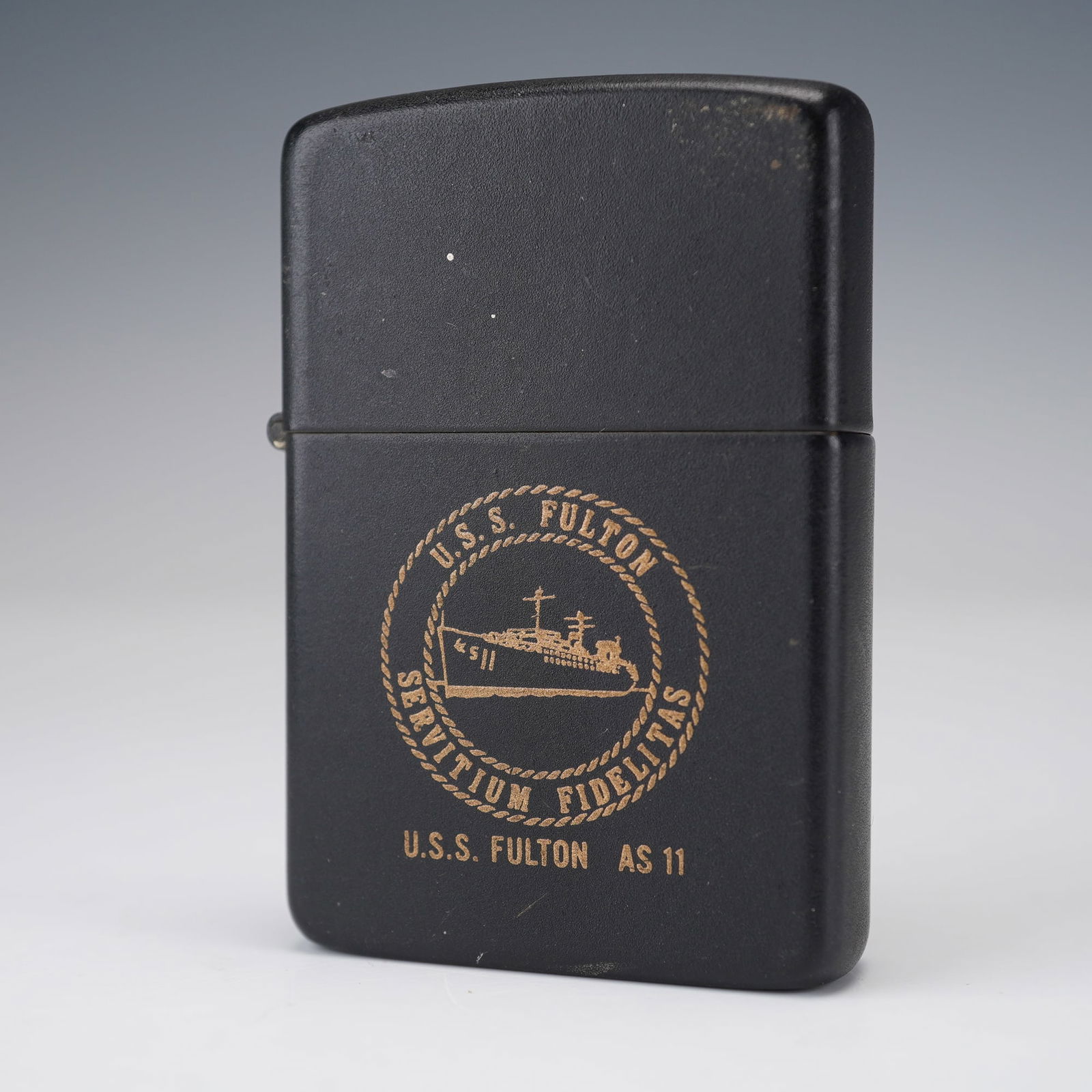 Vintage Zippo Lighter, U.S.S. Fulton AS 11: A vintage black matte Zippo lighter featuring the insignia and ship illustration of the U.S.S. Fulton AS 11, with the motto "Servitium Fidelitas" encircled by a rope border. The U.S.S. Fulton was a Un