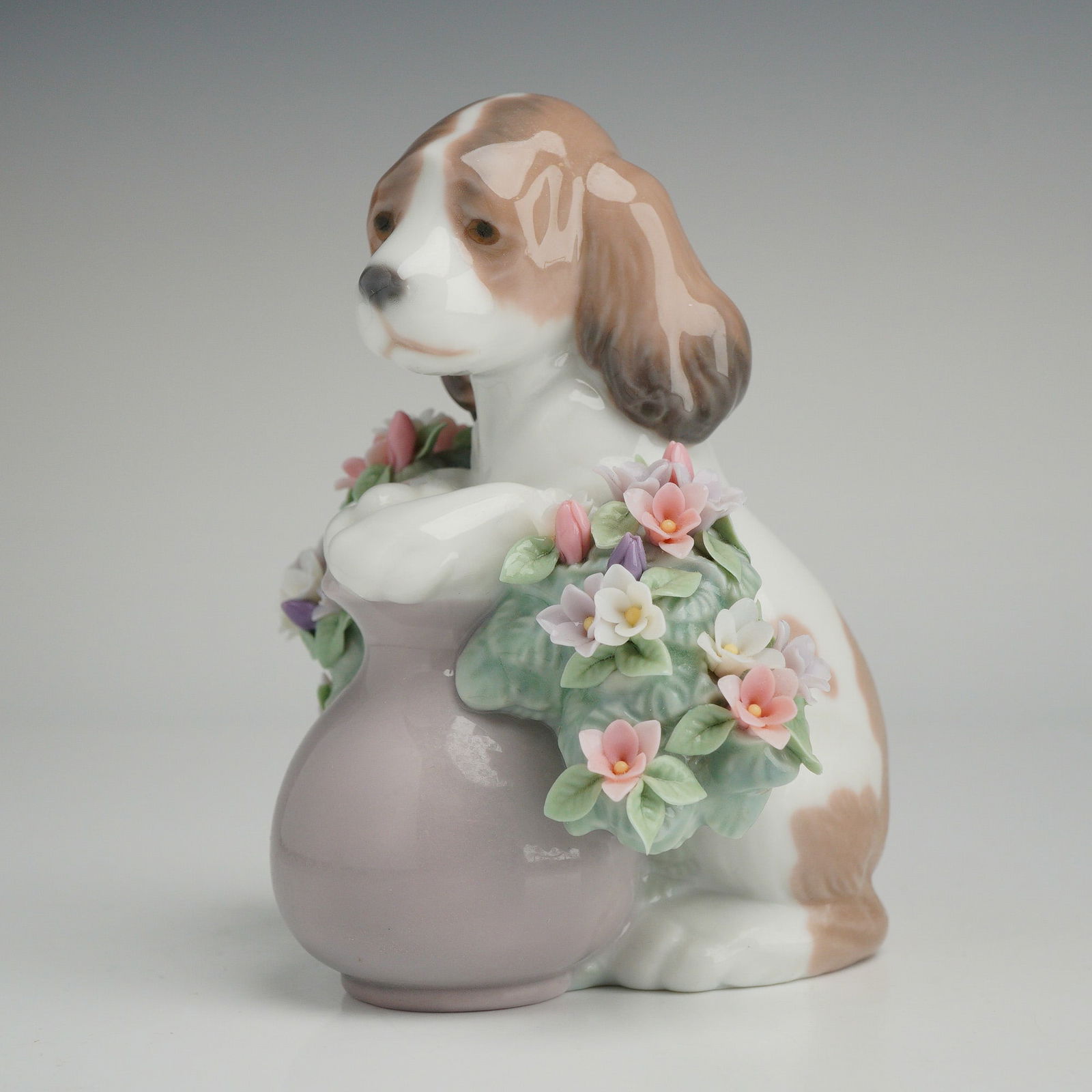 Lladro Figurine, Take Me Home 1006574: A Lladro Take Me Home glazed porcelain figurine, model number 1006574. The piece depicts a brown and white spaniel puppy with its front paws resting on a lavender-toned pot. The pot is adorned with a