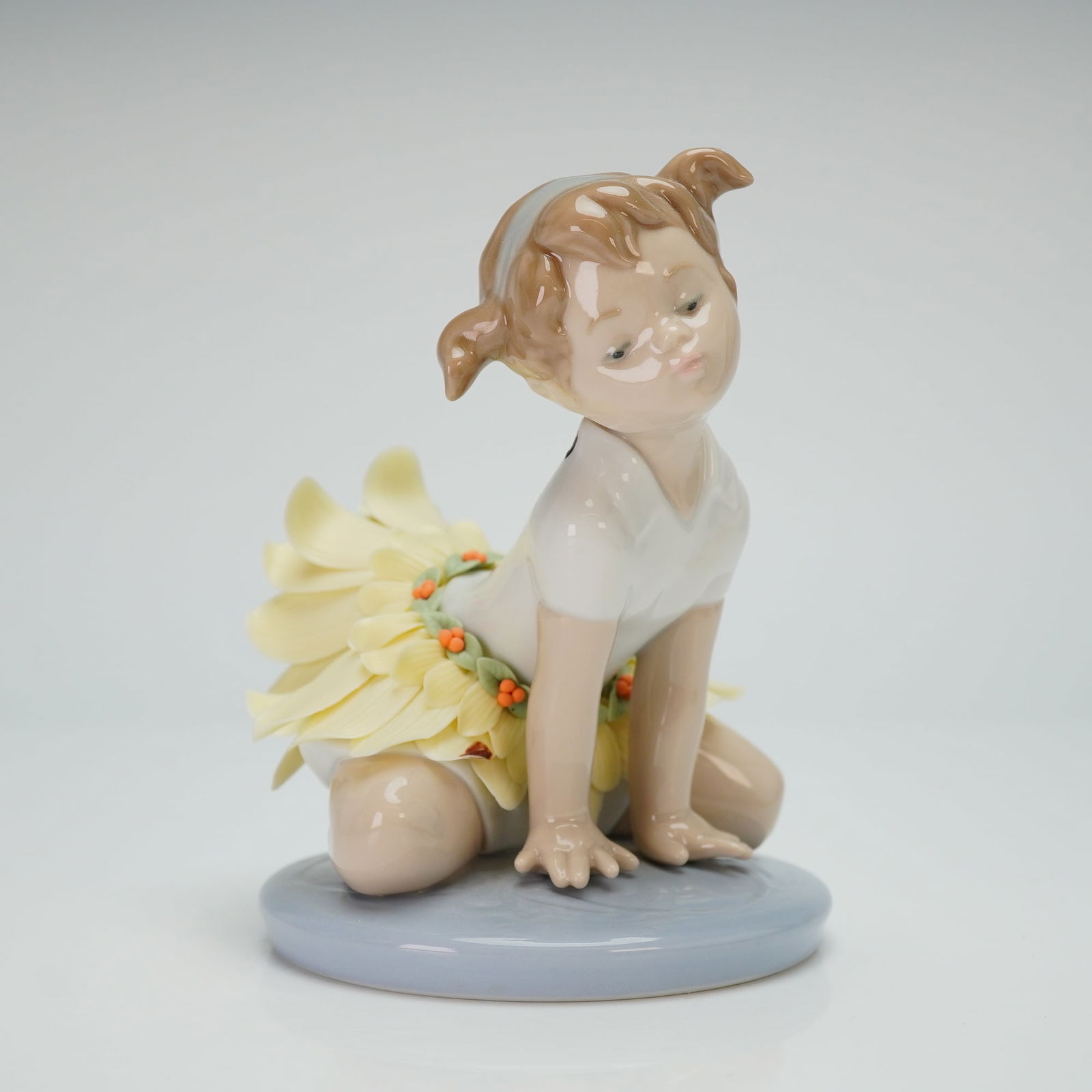 Lladro Porcelain Figurine Oopsy Daisy 01006691: The sculpture depicts a young girl with pigtails in a kneeling pose on a circular light blue base. She is dressed in a white leotard and a skirt composed of pale-yellow petals accented with a green vi