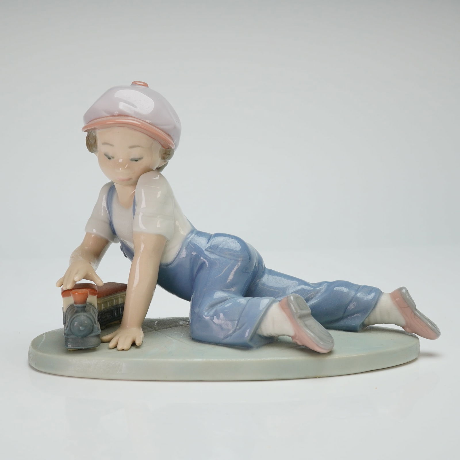 Lladro Porcelain Figurine All Aboard 01007619: A Lladro depicting a young boy in blue overalls and a pink-brimmed cap lying on his stomach while playing with a toy locomotive. The figure is executed in characteristic pale pastel tones with a high-
