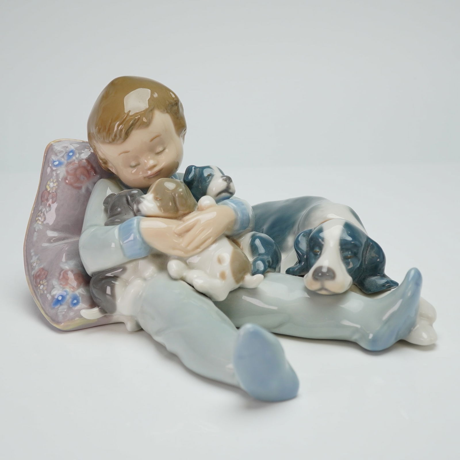 Lladro Porcelain Figurine Sweet Dreams 01001535: The piece depicts a seated young boy leaning against a lavender floral-patterned pillow, surrounded by several puppies. The figurine is executed in muted pastel tones of blue, gray, and brown with a h