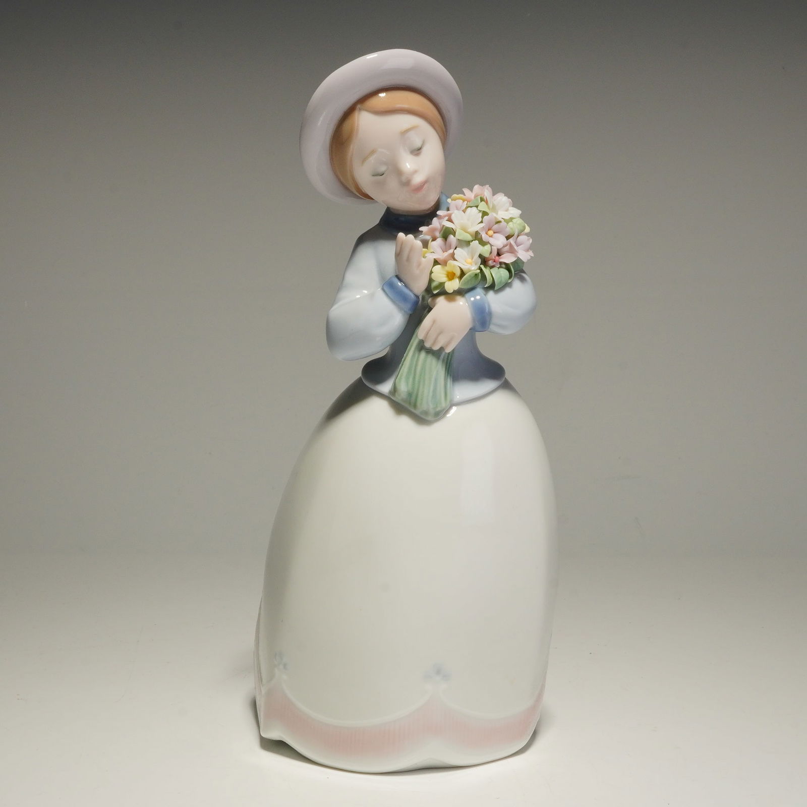 Lladro Porcelain Figurine, Blossom Time 01006869: The piece depicts a young woman in a wide-brimmed hat holding a bouquet of multicolored flowers. The figure is finished in a characteristic pastel palette, featuring a pale blue jacket and a long whit