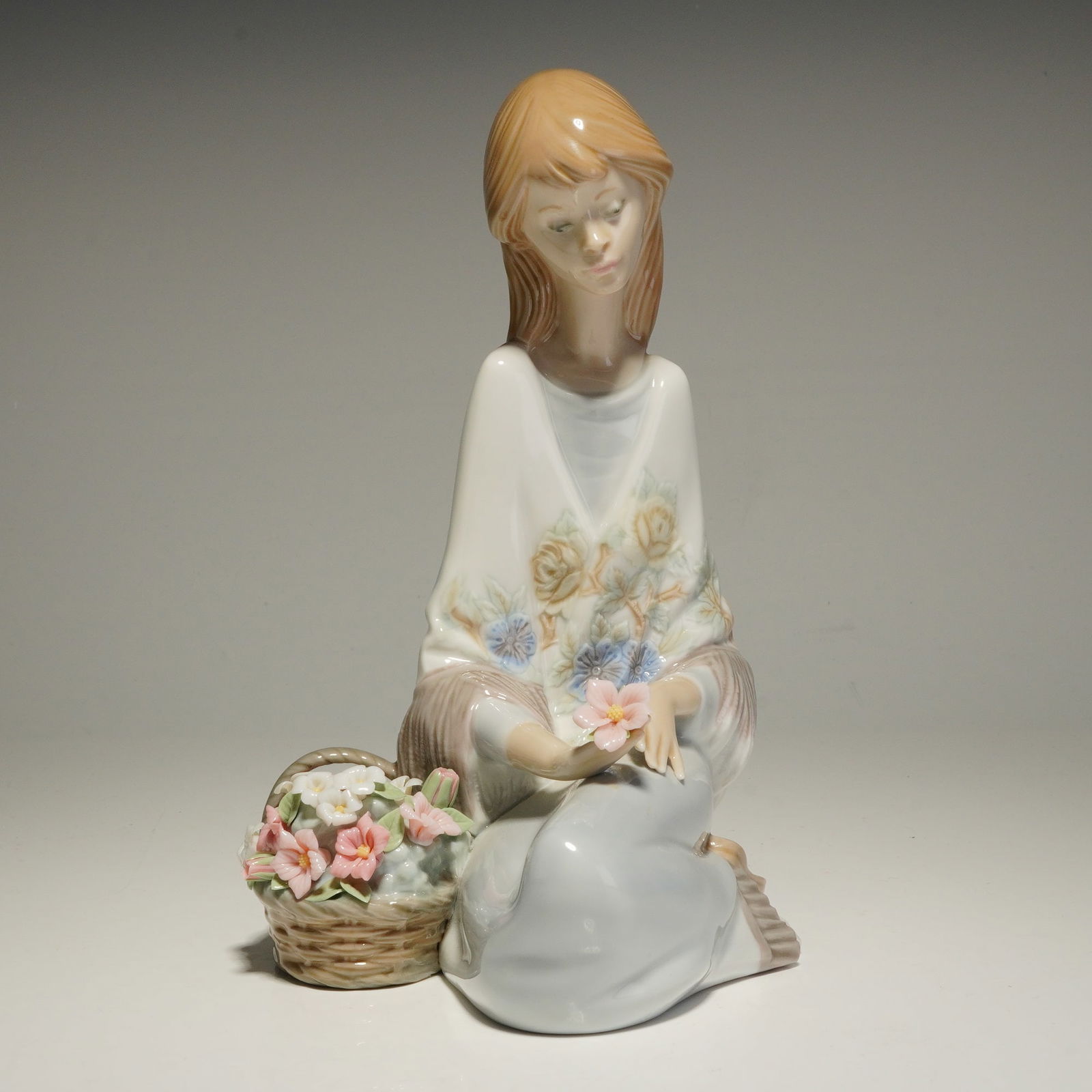 Lladro Porcelain Figurine, Flower Song 01007607: A Lladro glazed porcelain figurine originally issued for the Lladro Museum Collectors Society in 1988. The figurine depicts a young woman kneeling beside a basket of hand-modeled flowers while holding