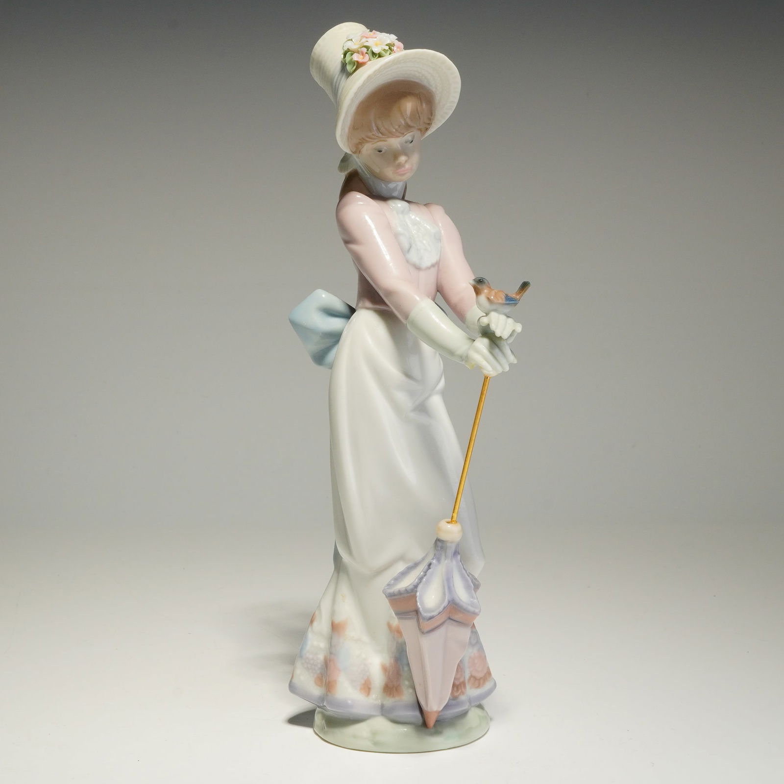 Lladro Porcelain Figurine, Garden Song 01007618: The figure depicts a young woman in period attire, wearing a wide-brimmed hat adorned with pastel-colored flowers. She is posed holding a small bird on her gloved hands and carries a closed parasol wi