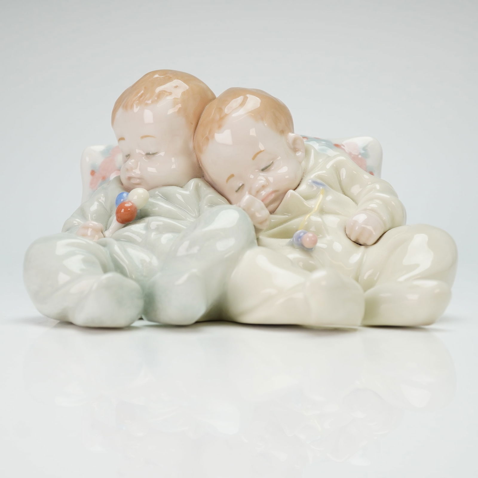 Lladro Porcelain Figurine, Little Dreamers 01005772: A Lladro porcelain figurine depicting two sleeping infants reclining against a large floral pillow. The figures are finished in a pastel palette, with one infant in pale blue pajamas and the other in