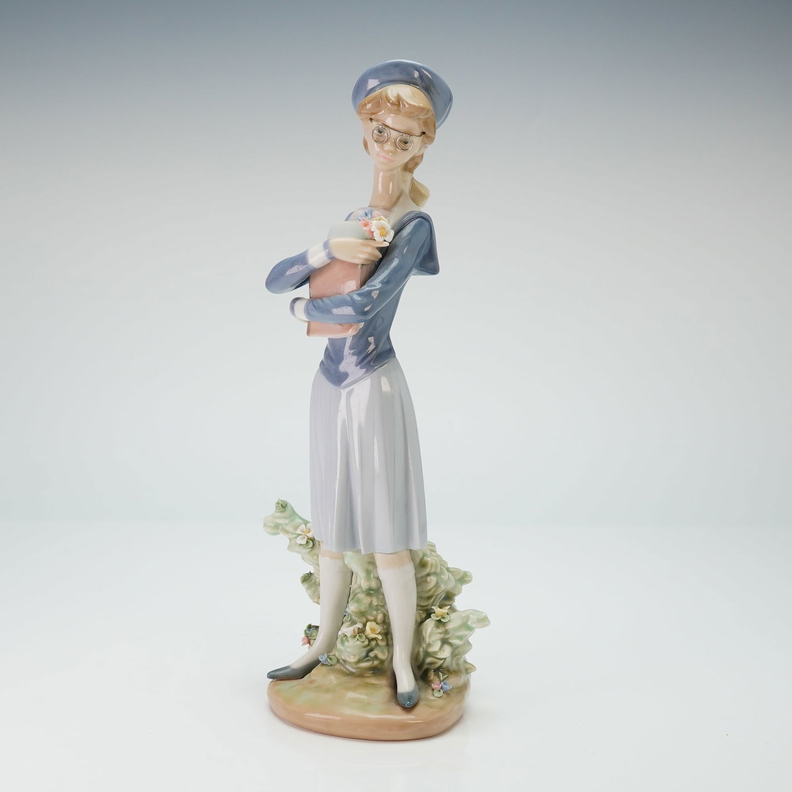 Lladro Porcelain Figurine, My First Class 01005708: Lladro depicting a young female student dressed in a blue and gray sailor-style school uniform. The figure is modeled wearing glasses and holding a stack of books with a small blossom. The figurine is