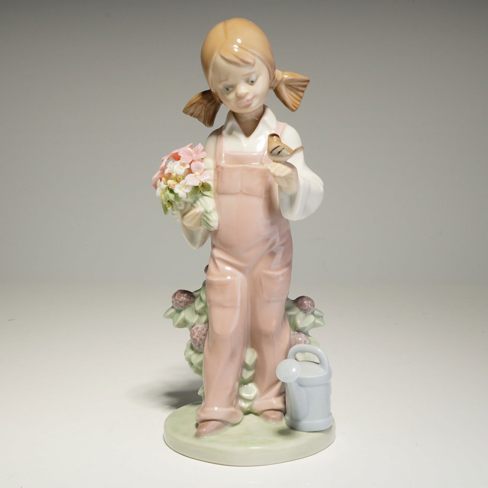 Lladro Porcelain Figurine, Spring Girl 01005217: The piece depicts a young girl with pigtails wearing pink overalls and a white long-sleeved shirt. She holds a cluster of pink and white flowers in her right hand, while a small brown bird perches on