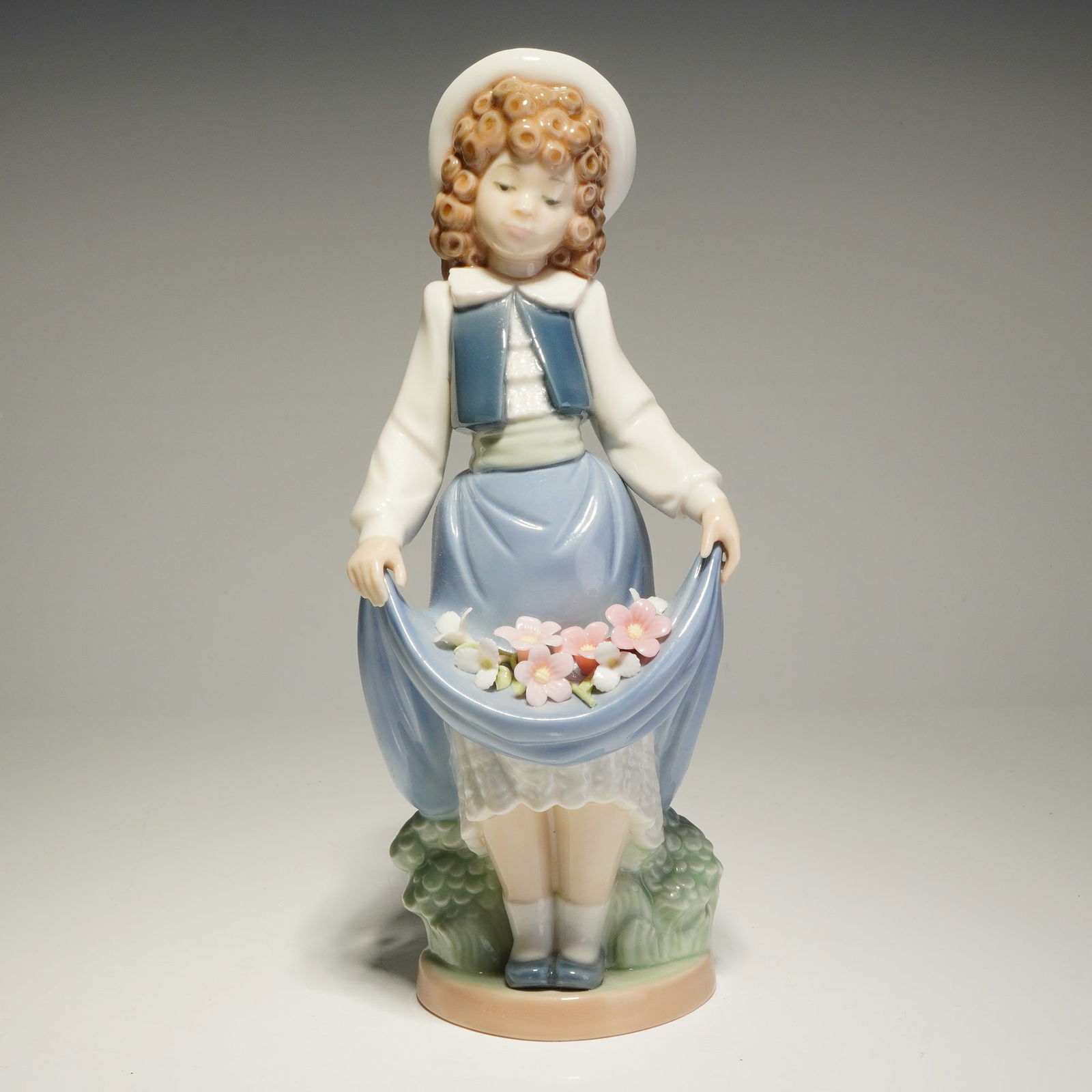 Nao by Lladro Porcelain Figurine The School Girl Number 1005: The figure depicts a young girl with curly brown hair wearing a white hat, a blue vest, and a blue skirt. She is shown holding a cluster of pink and white flowers within the fold of her skirt. The pie