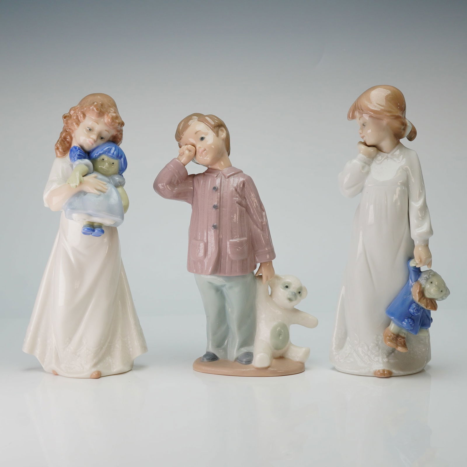 3pc Nao by Lladro Porcelain Figurines: A trio of Nao by Lladro glazed porcelain figurines depicting children in sleepwear. The collection includes the model We're Sleepy 1107, featuring a girl in a nightgown holding a doll in a blue coat,