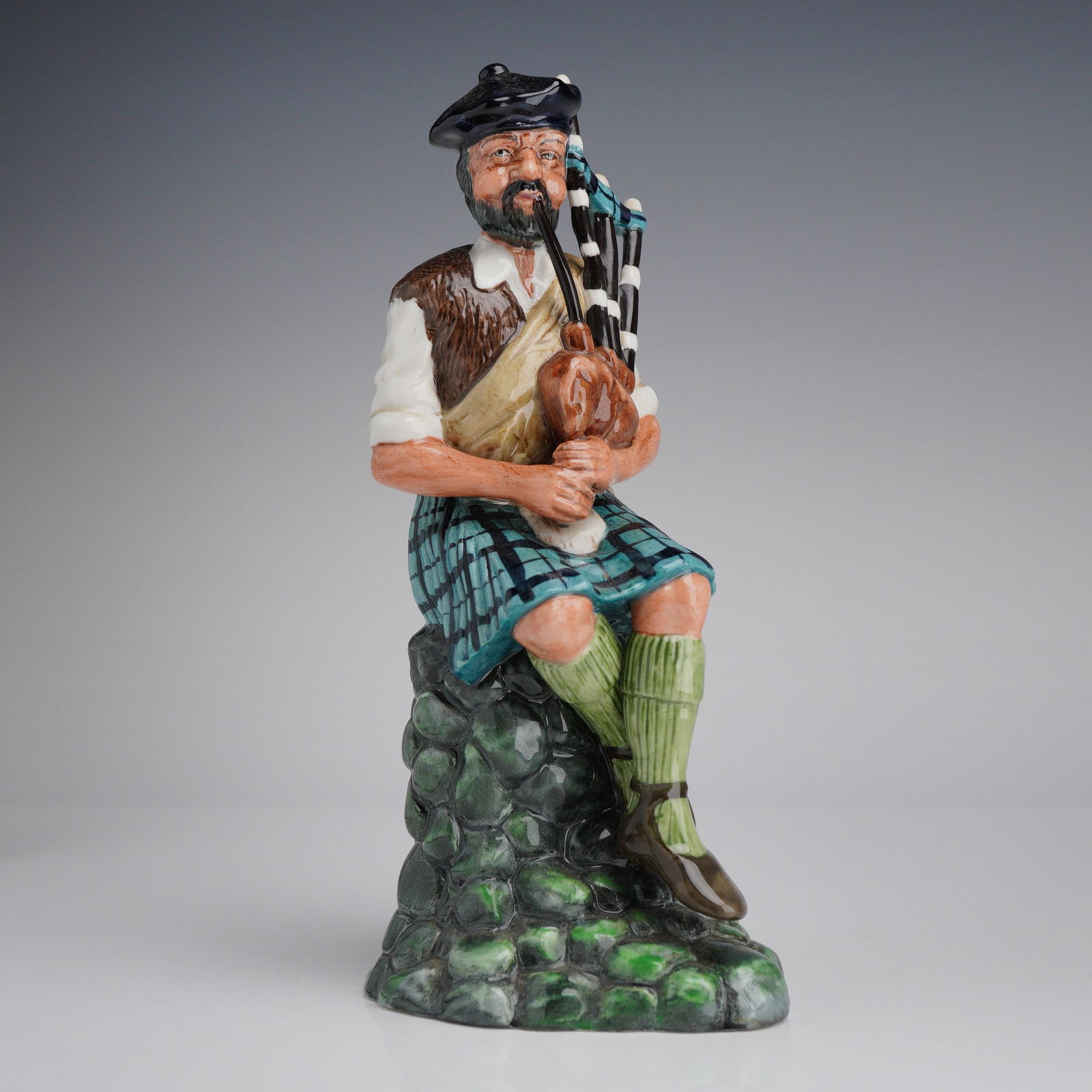 Royal Doulton Figurine The Piper HN2907: The figure depicts a bearded man in traditional Scottish attire seated upon a stone wall. The subject is modeled wearing a blue and green tartan kilt, a brown waistcoat over a white shirt, green hose,