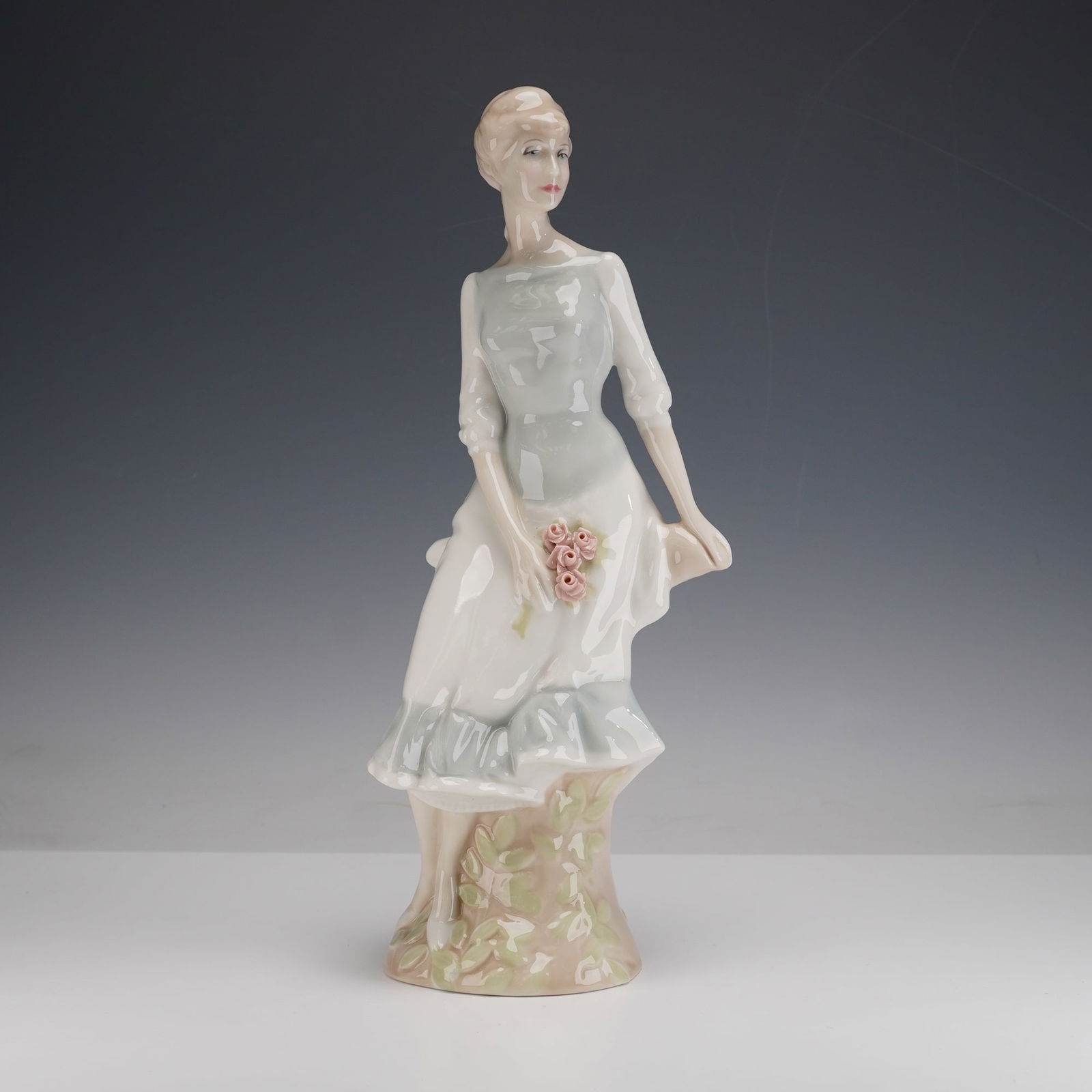 Royal Doulton Reflection Collection Figurine, Rose Arbour HN3145: This figurine depicts a woman seated on a tree stump while holding a small bouquet of pink roses. She wears a long pastel dress with molded folds, and the tree trunk base has foliage details. Royal Do