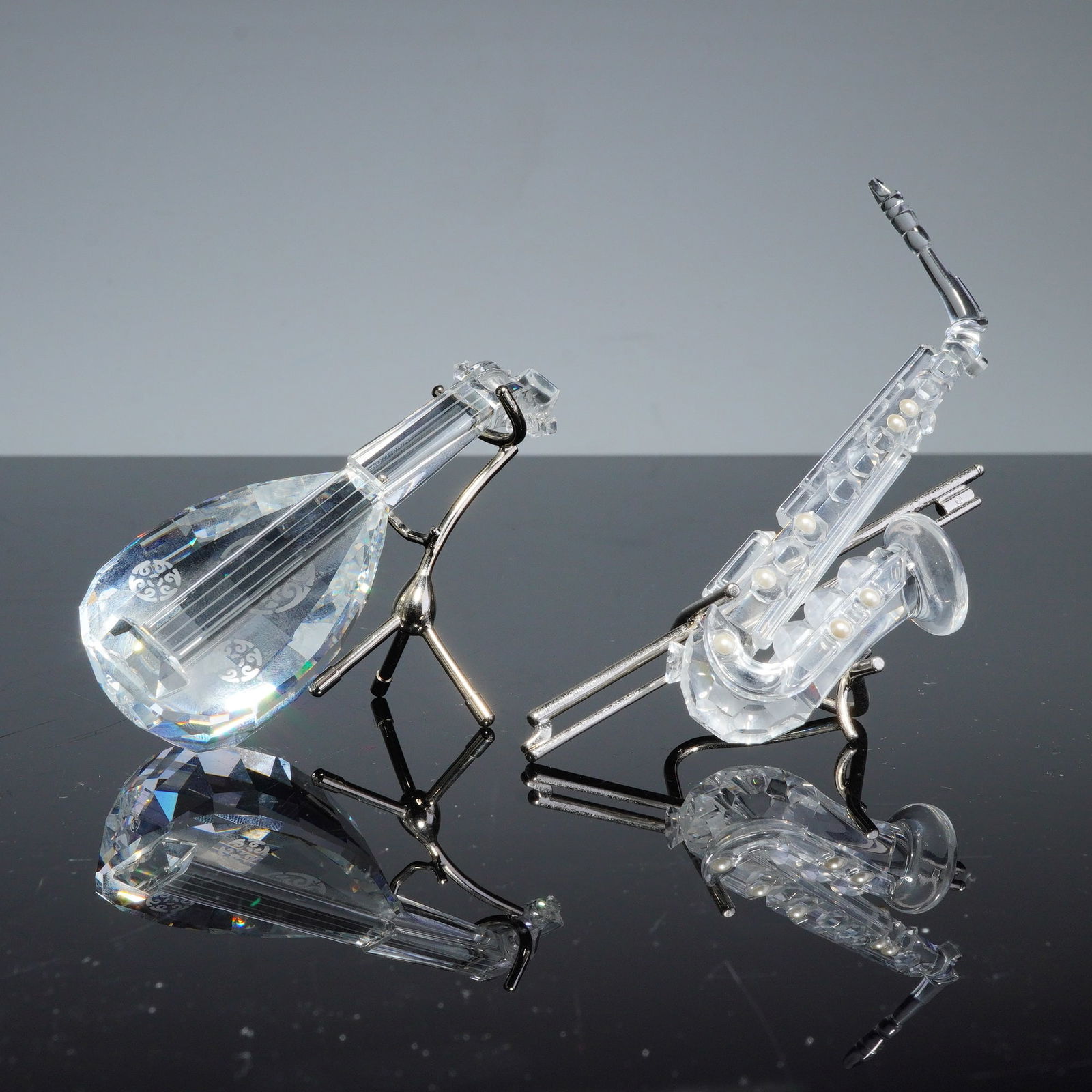 2pc Swarovski Silver Crystal Figurines, Lute and Saxophone: A pair of Swarovski faceted crystal musical instrument figurines from the Silver Crystal collection, comprising a lute and a saxophone. Each piece is crafted from clear lead crystal with precision-cut