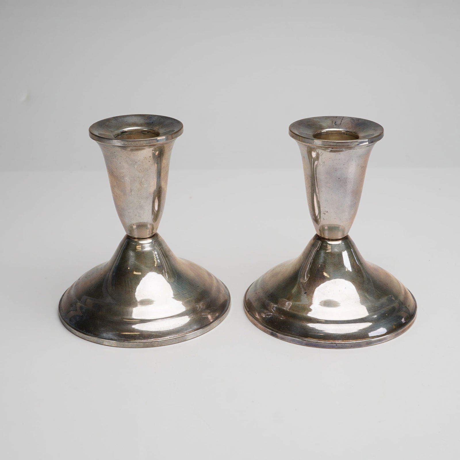 Pair of Duchin Creation Sterling Silver Weighted Candleholders: Each candlestick features a simple, trumpet-shaped cup on a short stem that transitions into a wide, stepped, and domed circular base. The undersides are stamped with the manufacturer hallmark depicti