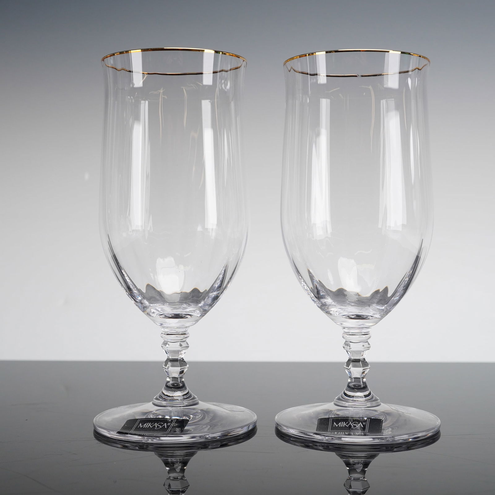 A Pair of Mikasa Crystal Iced Tea Glasses in the Sonata Pattern: A pair of Mikasa crystal iced tea glasses in the Sonata pattern. Each glass features a clear tapered bowl decorated with vertical panel cuts at the base and a thin gold-gilt rim. The bowls are support