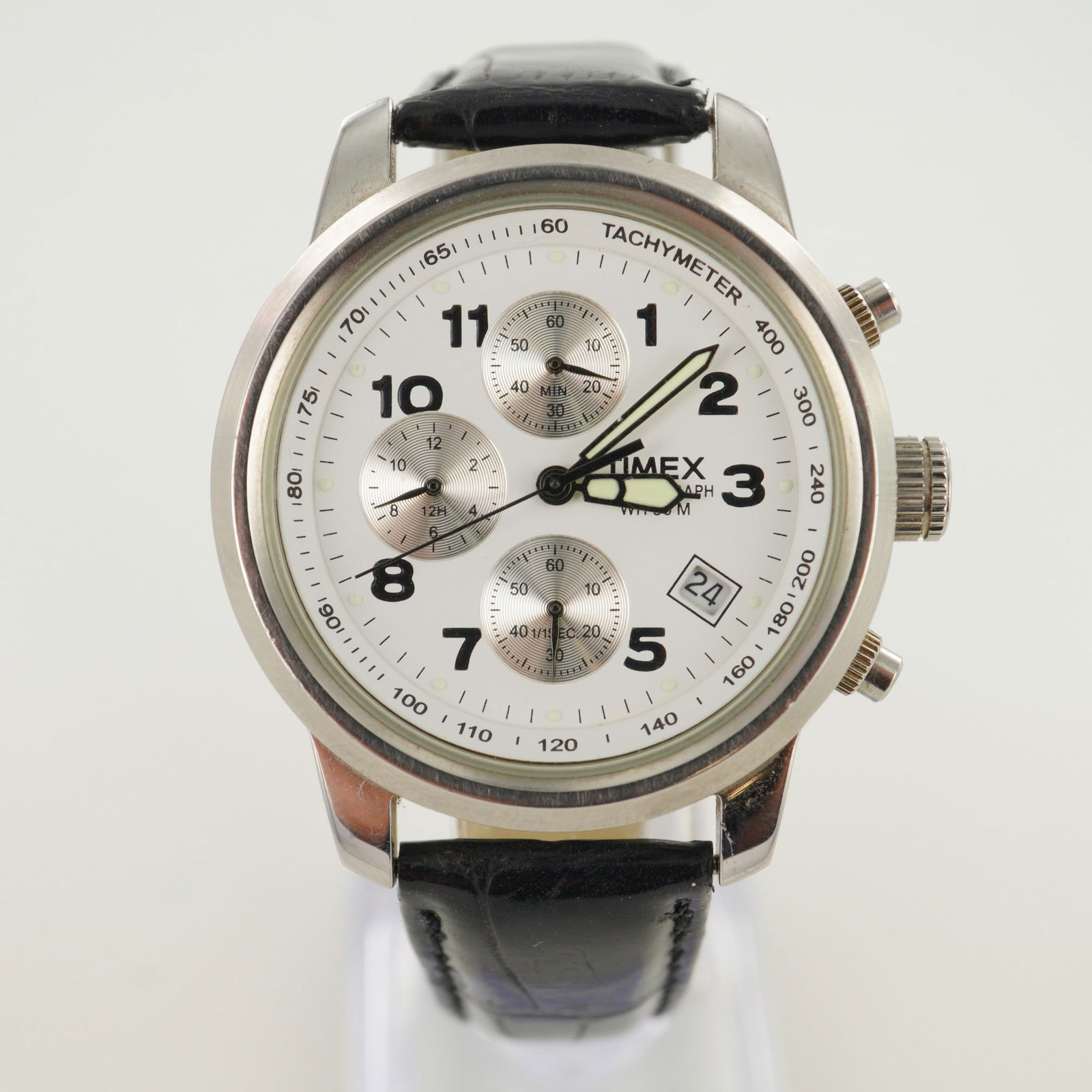 Timex Chronograph WR 50 M Wrist Watch: The watch features a white dial with three subdials for chronograph functions, a date window between the 4 and 5 o'clock positions, black Arabic numeral hour markers, a tachymeter scale around the out