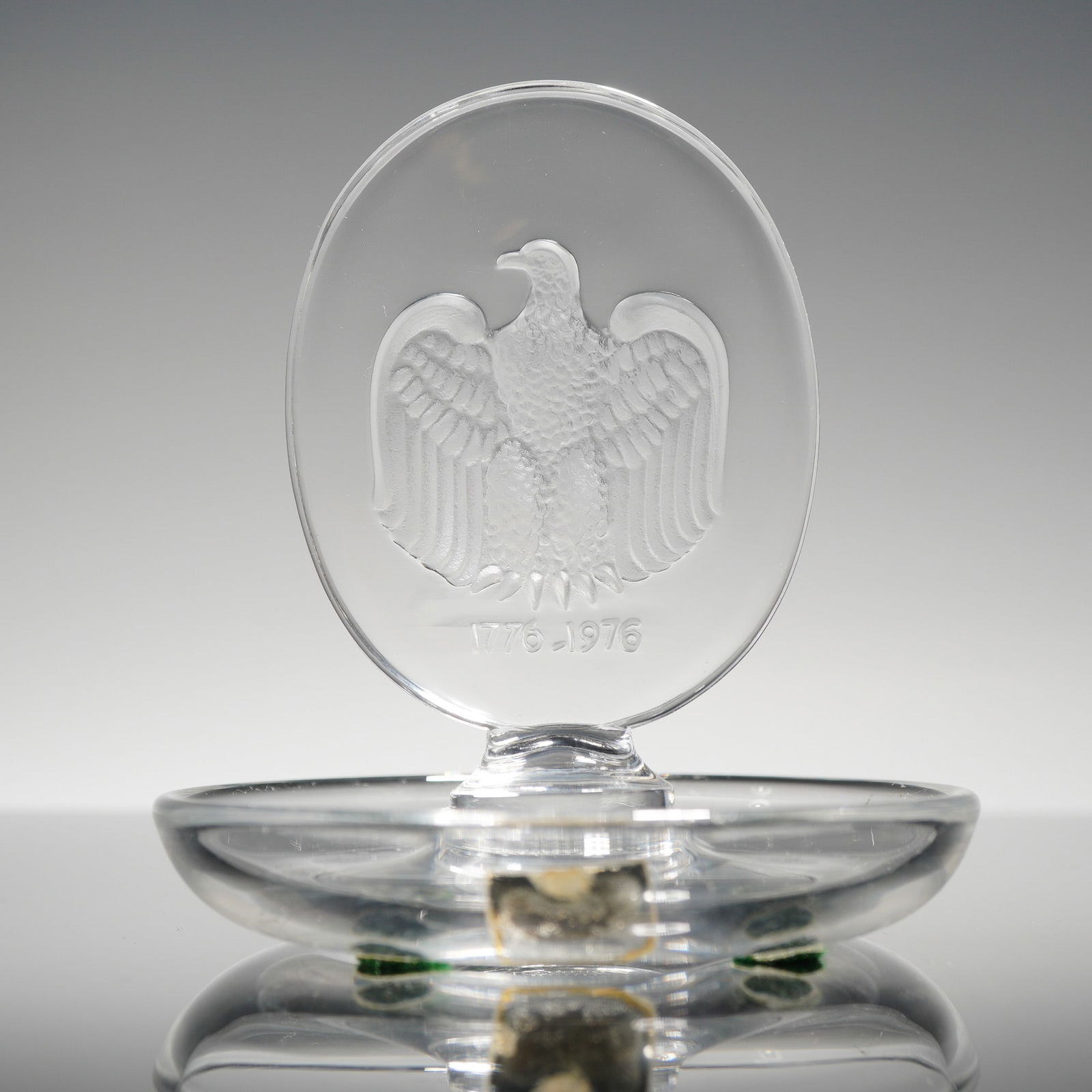 Lalique Round American Bicentennial Pin Tray: A Lalique France clear and frosted glass pin tray in the Aigle pattern, produced circa 1976 to commemorate the American Bicentennial. The item features a vertical oval medallion with a frosted intagli