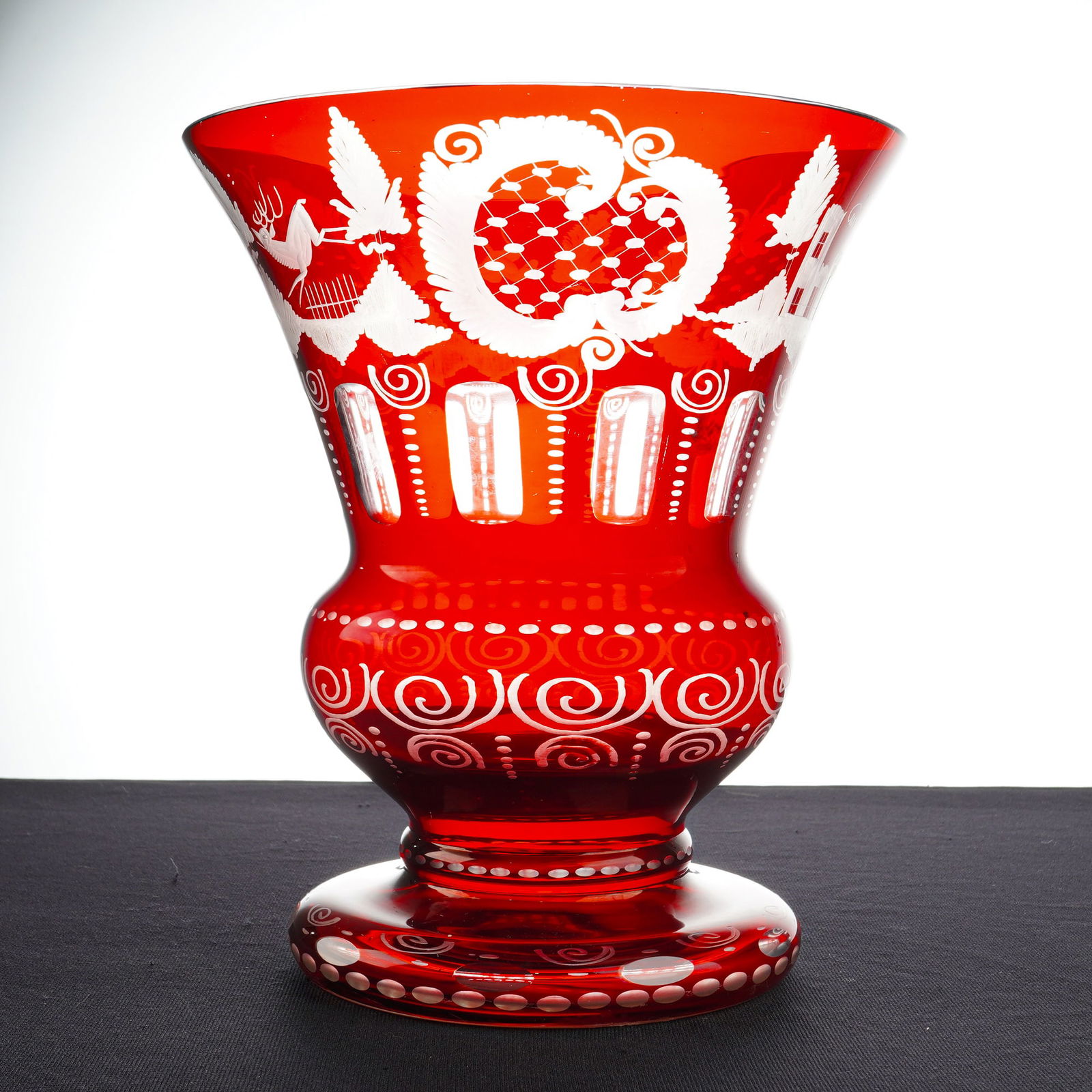 Bohemian Ruby Red Cut to Clear Glass Vase: A red cut to clear glass vase featuring a wide, flared rim above a bulbous midsection and a pedestal base. The upper body is decorated with detailed wheel-etched motifs including a stag, a manor house