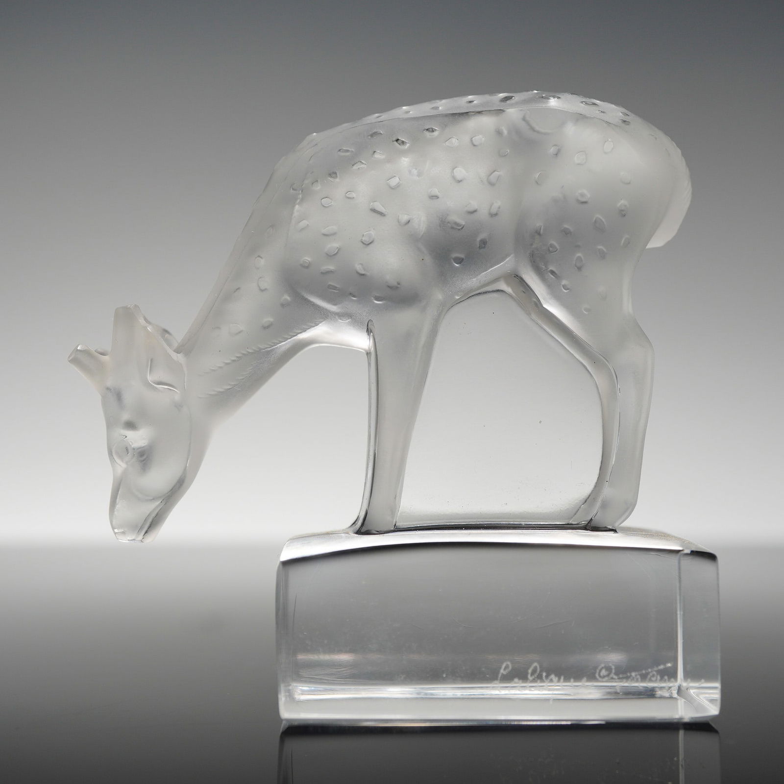 Lalique France Frosted Crystal Paperweight, Fawn: The piece is constructed of molded glass with a frosted satin-finished body featuring circular spots. The figure is shown in a downward grazing pose atop an integral, polished clear rectangular base.