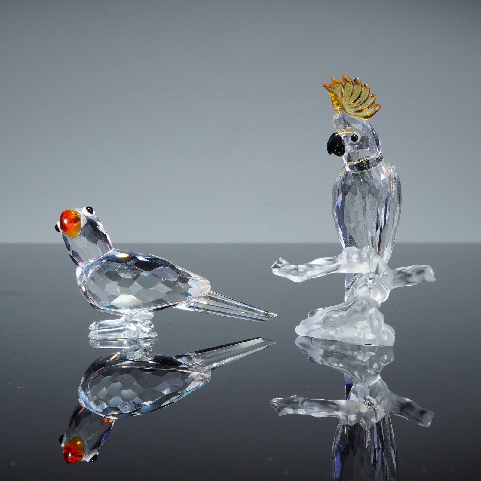 2pc Swarovski Crystal Bird Figurines: This pair of Swarovski crystal figurines includes the Cockatoo, model number 261635, and a parrot with orange-red accents. Each piece is crafted from faceted clear lead crystal. The cockatoo features
