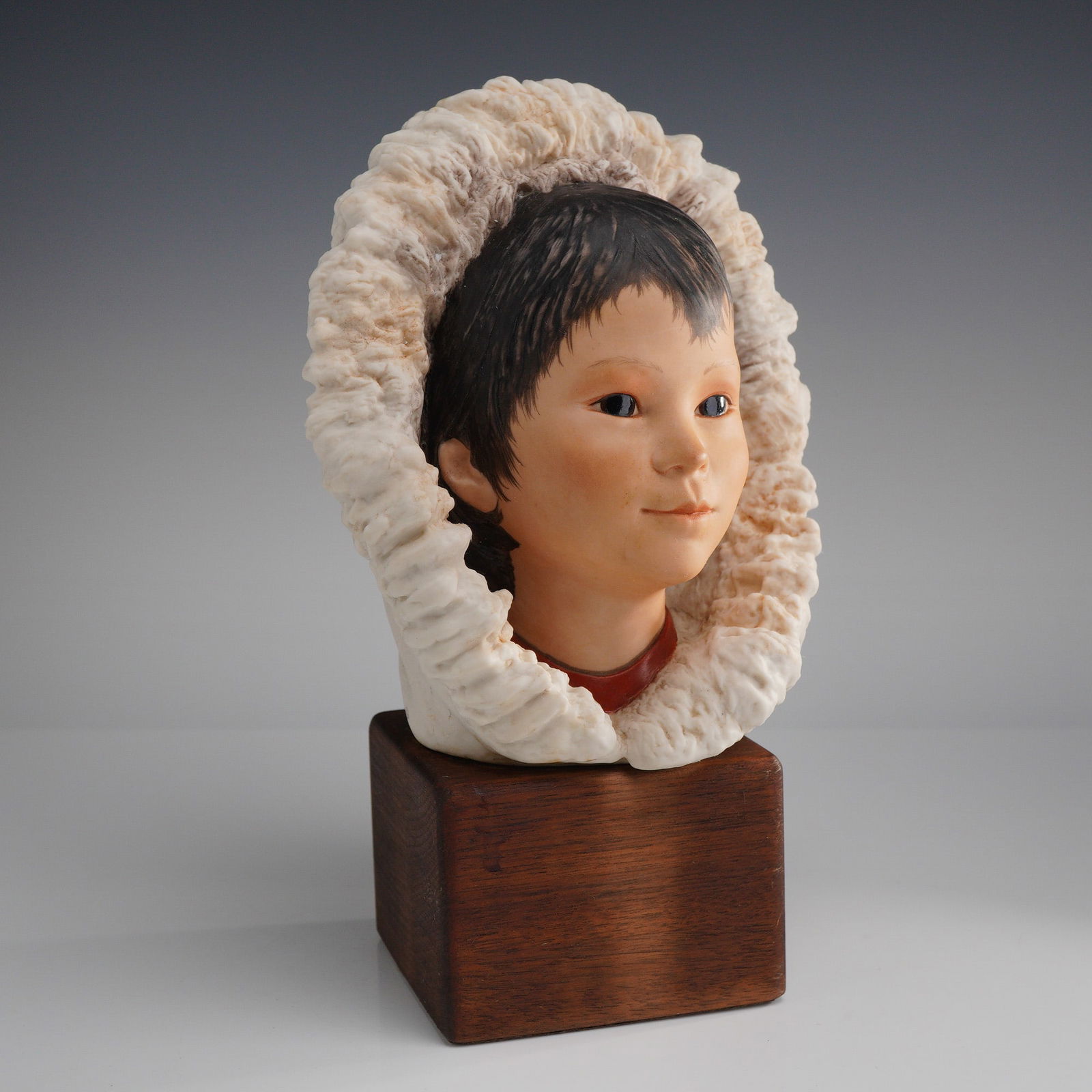 Cybis Bisque Porcelain Eskimo Child Bust, Snow Bunting: The figure is shown wearing a hooded garment with a wide, highly textured off-white ruff. The face is realistically painted with dark eyes and hair. The porcelain bust is mounted on a square, dark woo