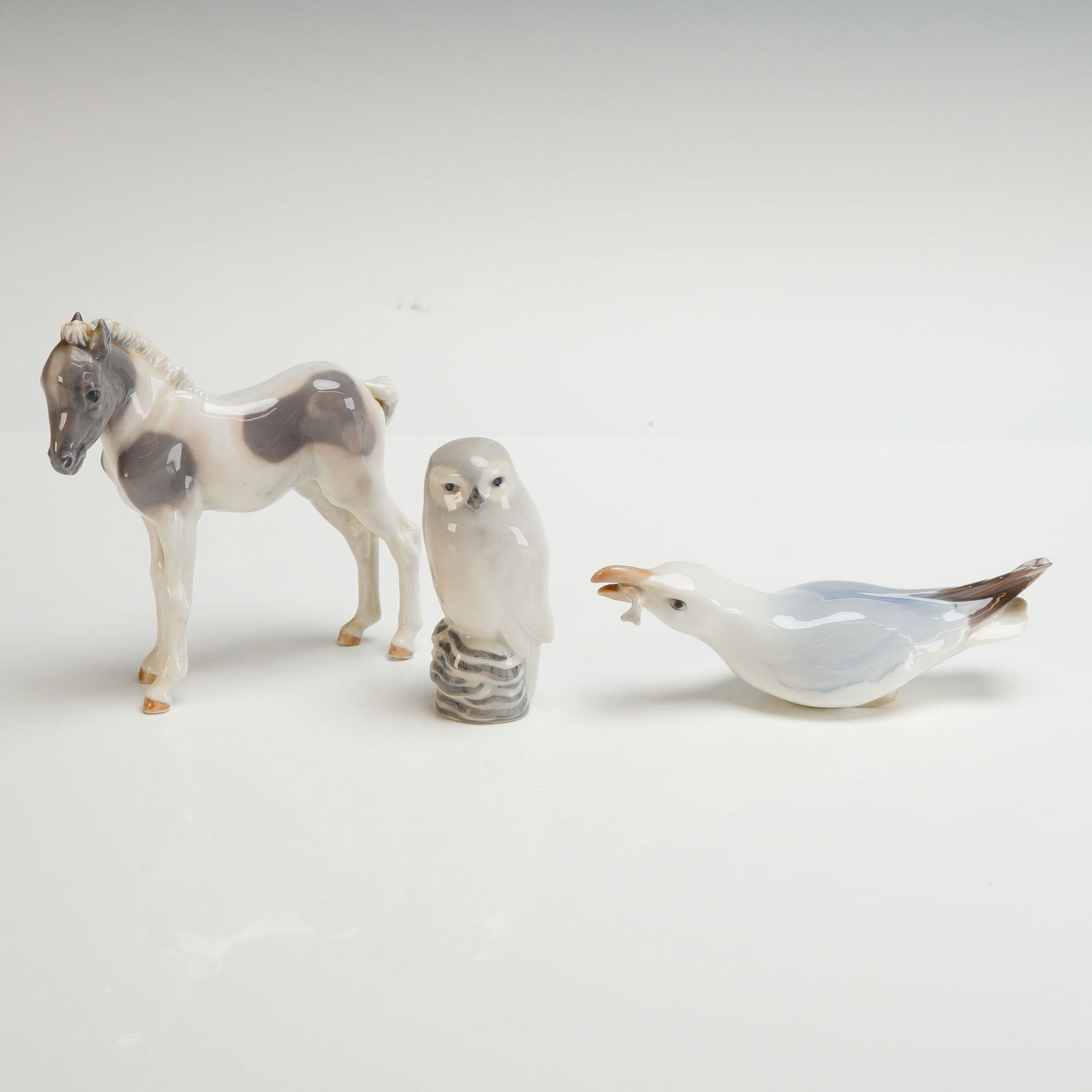 3pc Royal Copenhagen and Bing & Grondahl Animal Figurines: A collection of three Danish glazed porcelain figurines including a foal, an owl, and a seagull with a fish. Each piece is rendered in a naturalist style with muted underglaze tones and a high-gloss f
