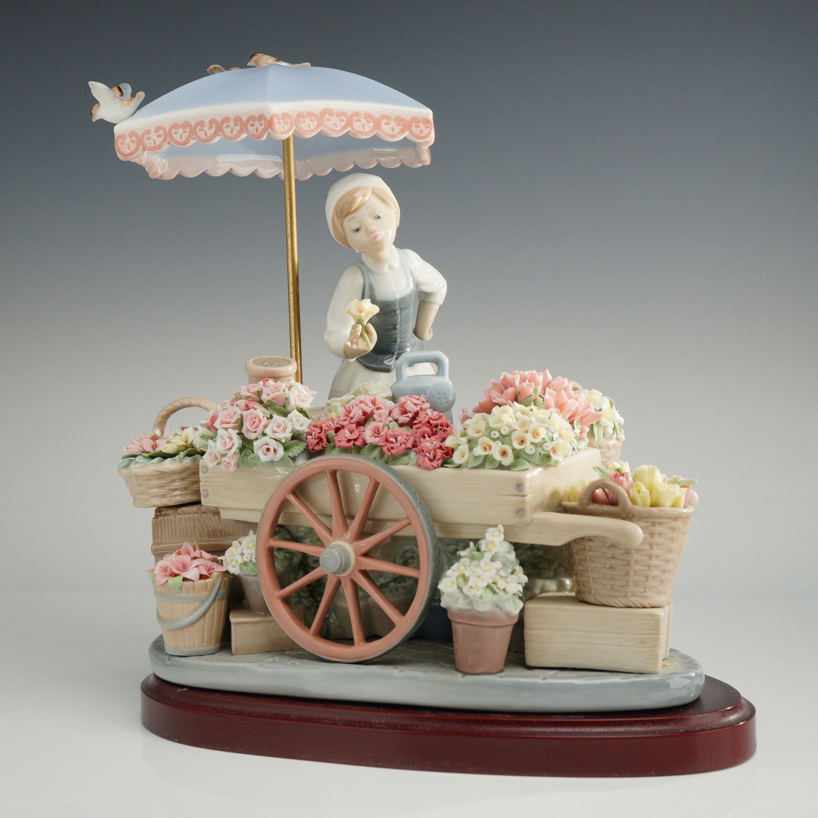 Lladro Flowers of the Season Porcelain Figurine 1001454: A Lladro glazed porcelain sculpture titled Flowers of the Season, model number 1001454. The figurine depicts a young female florist standing behind a flower cart laden with numerous hand-formed floral