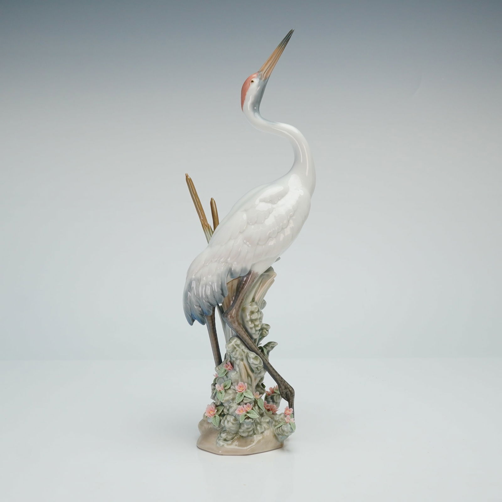 Lladro Porcelain Figurine Dancing Crane 01001614: This piece depicts a single slender white crane positioned amidst tall reeds and delicate pink floral accents. The figurine is executed in a traditional pastel palette and is mounted on an integral ba