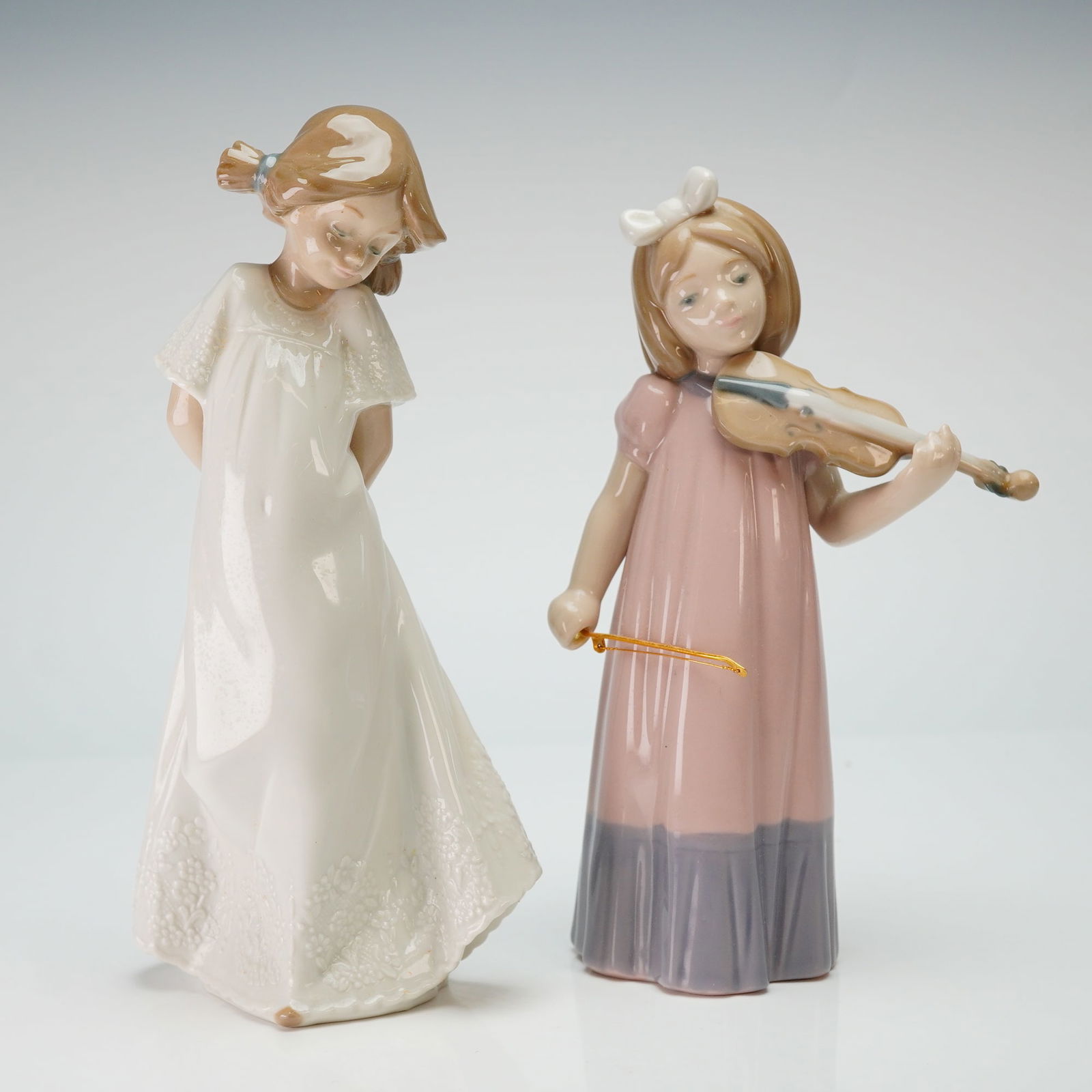 2pc Lladro Figurines, Girl with Violin and Girl in White Dress: Two Nao by Lladro glazed porcelain figurines. The group includes a figure of a girl playing the violin, identified as Nina Violin (Model 1034), and a figure of a girl in a long white dress with floral