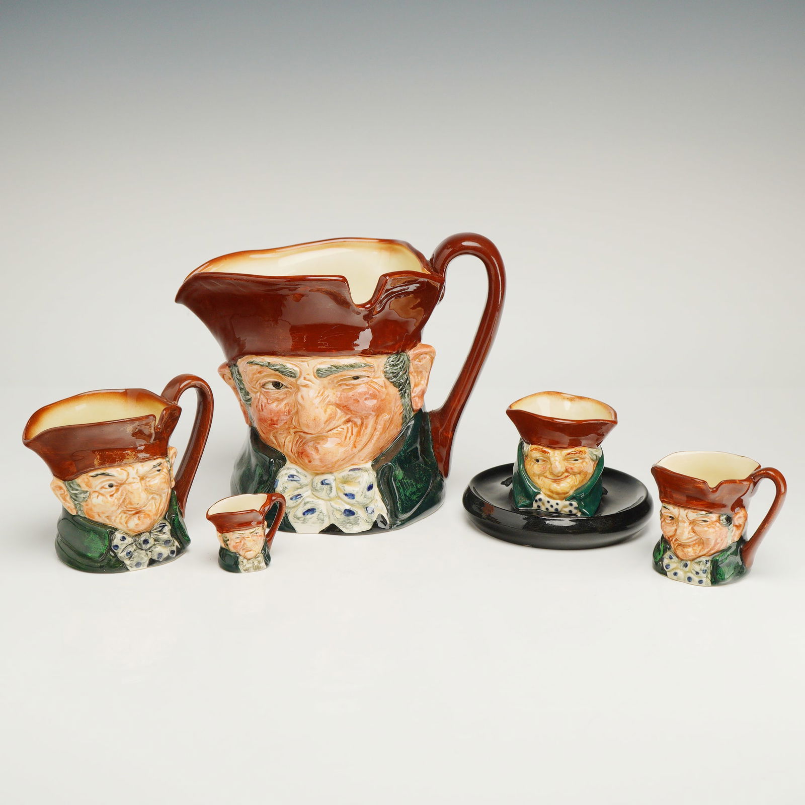 5pc Royal Doulton Character Jugs Old Charley: A 5pc collection of Royal Doulton glazed ceramic character jugs featuring the Old Charley in a variety of sizes and shapes. The Old Charley pieces are depicted in a brown tricorn hat and green coat wi