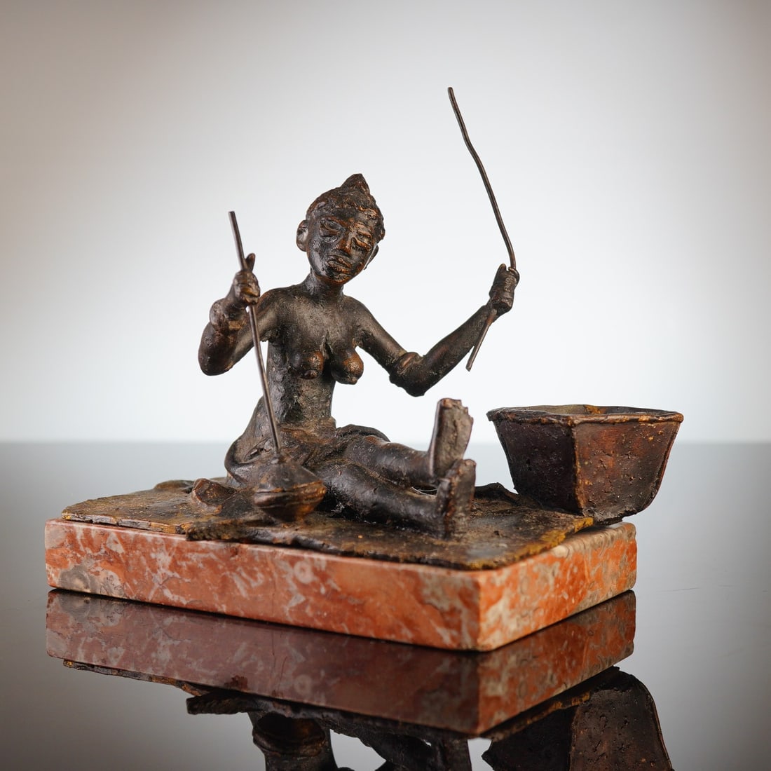 African Bronze Figurine Mounted on a Stone Base: This cast bronze figurine of African influence depicts a seated, bare-chested figure holding two thin rods. The figure is positioned on a textured metal plinth adjacent to a square-shaped vessel. The