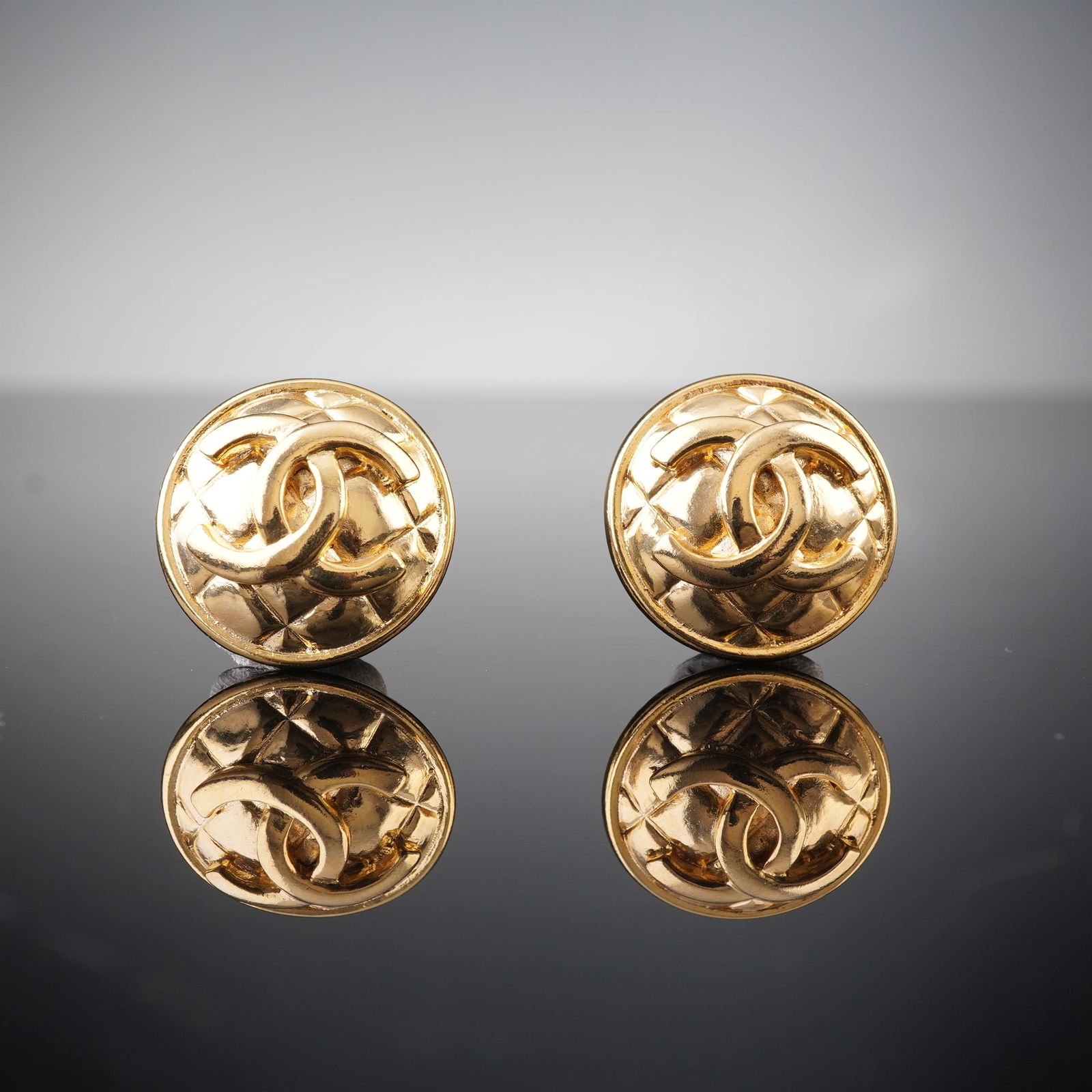 A Pair of CHANEL Gold-Tone CC Quilted Round Buttons: A pair of CHANEL gold-tone CC quilted round button. Each button prominently displays an interlocking double 'C' logo at its center, set against a raised, quilted or matelasse pattern. Dimensions