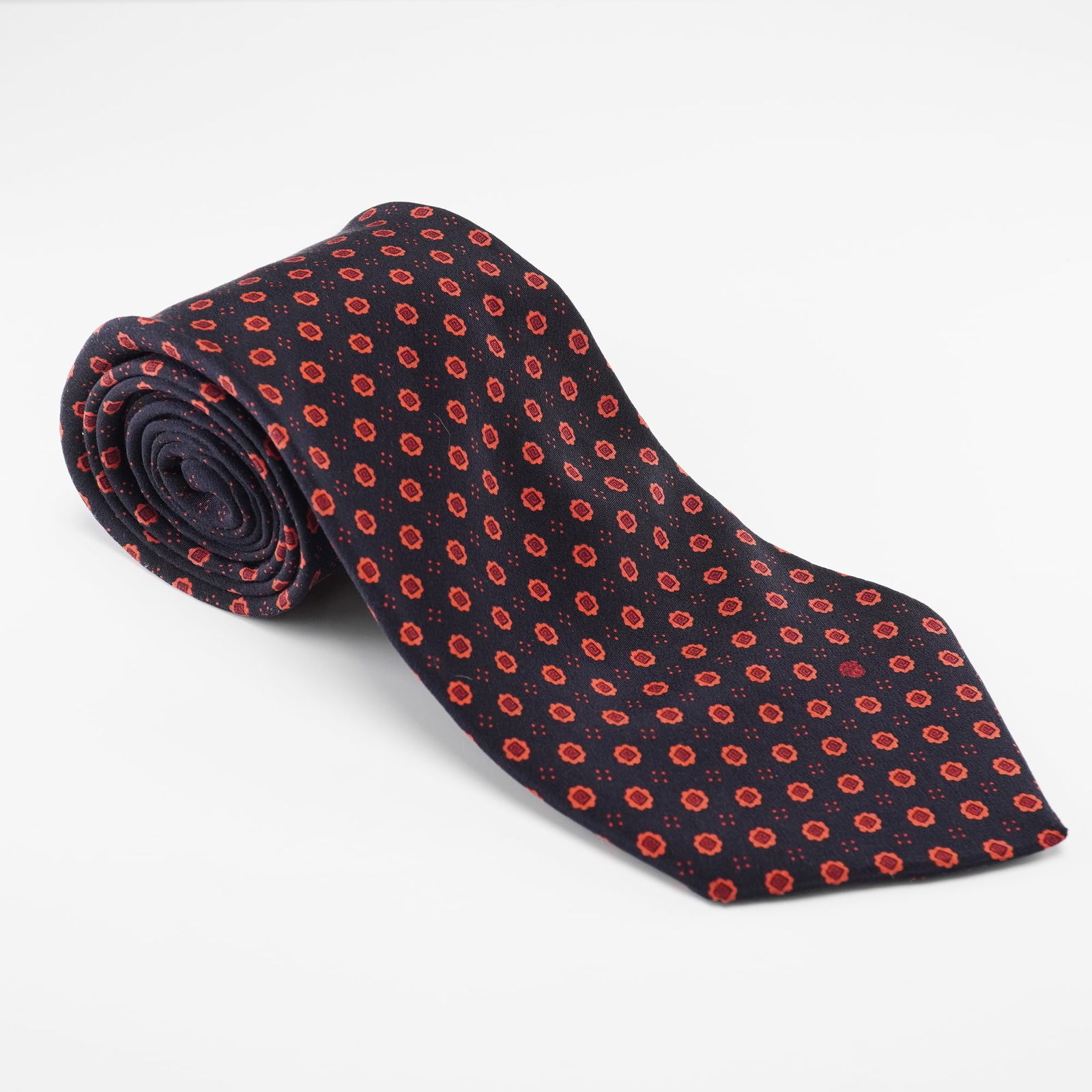 A Versace Silk Necktie, Navy Blue with Red Floral Motif, Made in Italy: A Versace silk necktie, crafted in dark navy blue silk, featuring a repeating pattern of small, vibrant red, four-petaled floral or geometric motifs. The fabric exhibits a slight sheen. A white label