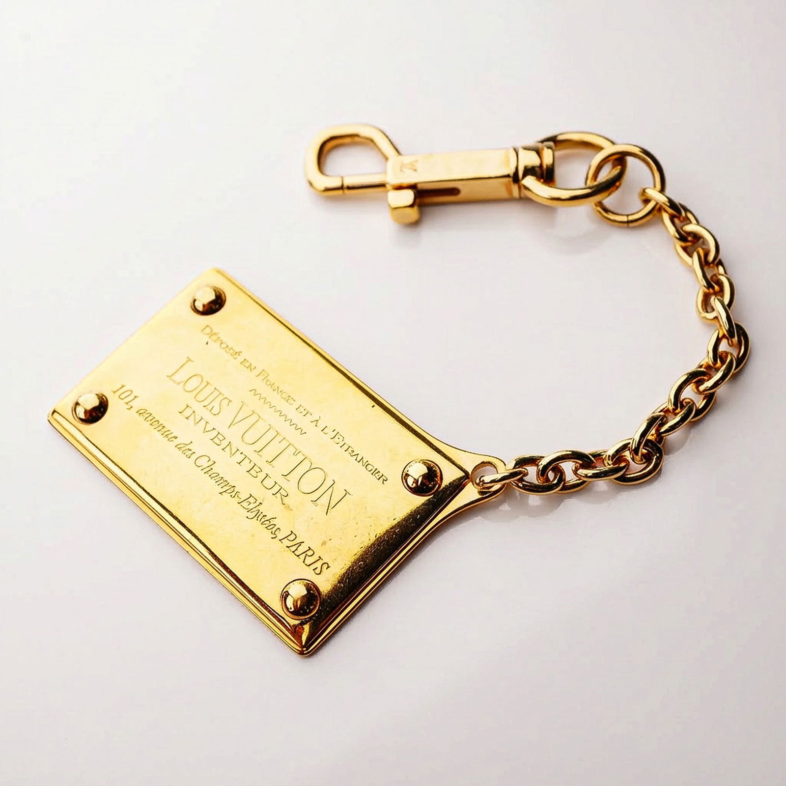A Louis Vuitton Gold-Tone Plaque Bag Charm: A single Louis Vuitton gold-tone bag accessory constructed as a key holder or bag charm. The item features a trapezoidal plaque secured by four visible rivets, connected to a short chain of thick link