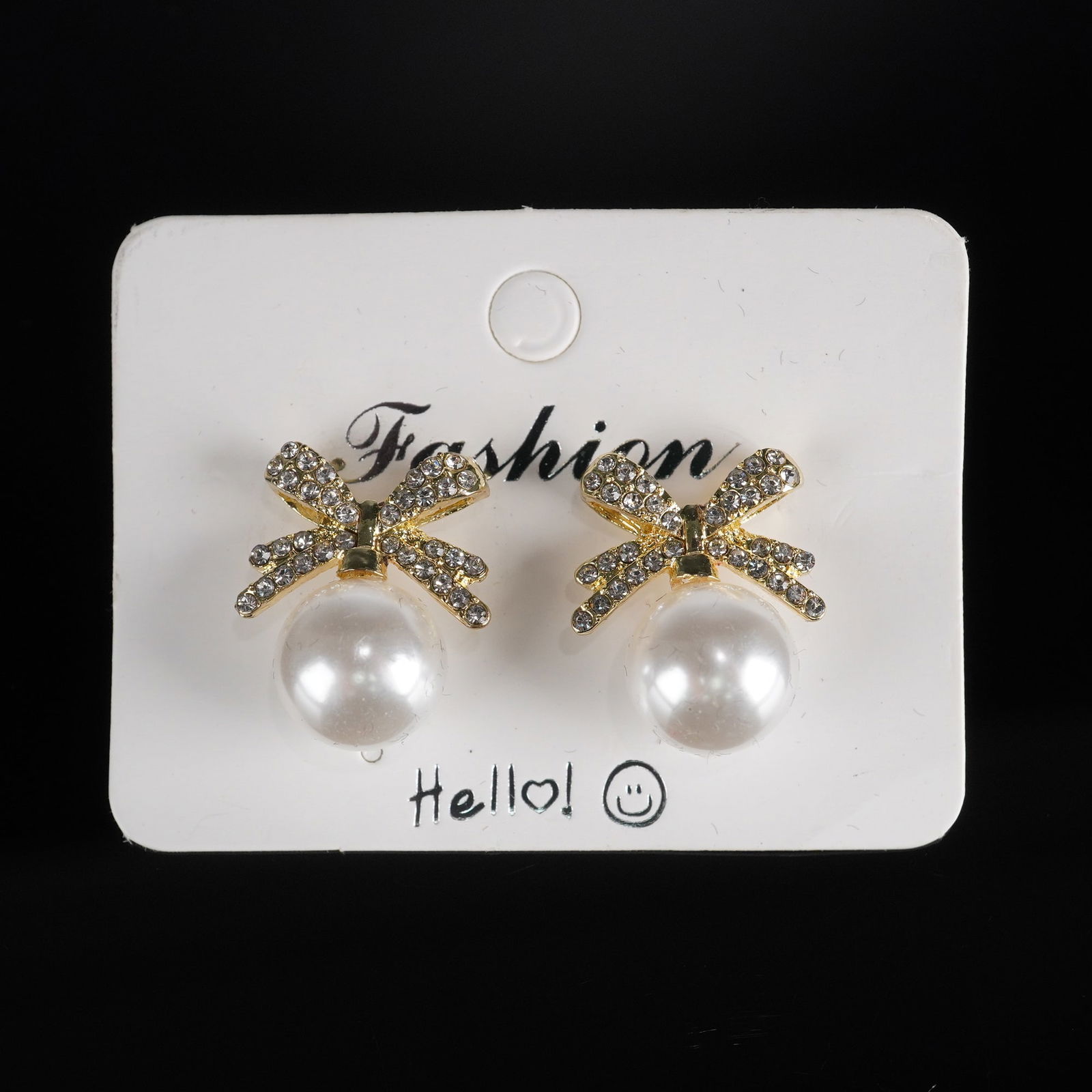 Pair of Modern Fashion Gold-Tone Metal and Faux Pearl Bow Stud Earrings: A pair of modern fashion stud earrings, each featuring a gold-tone metal bow design, pave-set with numerous clear, round faceted stones. Suspended from the bow is a single spherical white faux pearl.