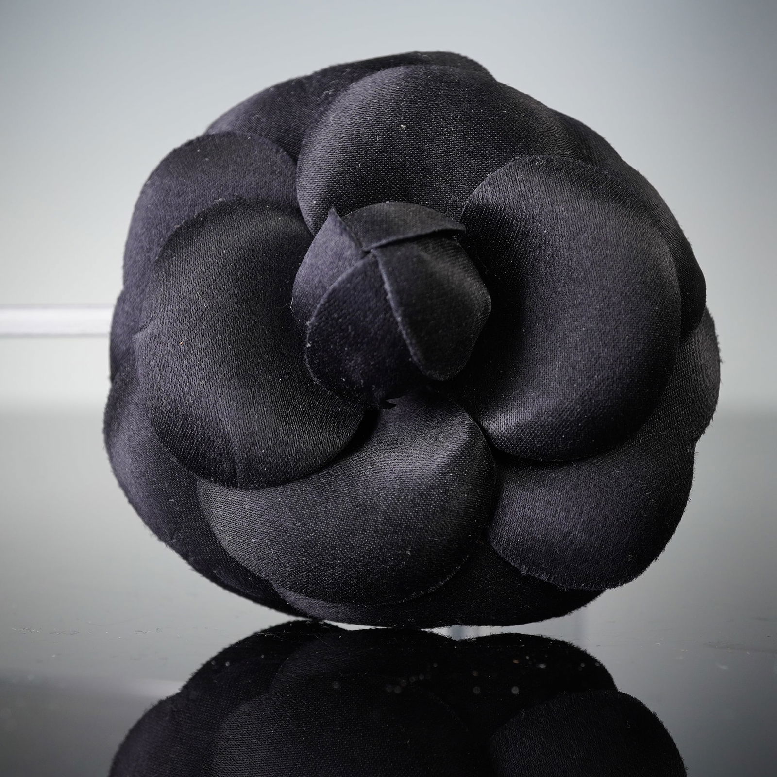 A Chanel Black Fabric Camellia Brooch: A Chanel Black Fabric Camellia Brooch, crafted from a satin-like fabric forming multiple layered petals. The reverse features a functional gold-tone metal safety pin clasp mechanism. It is accompanied