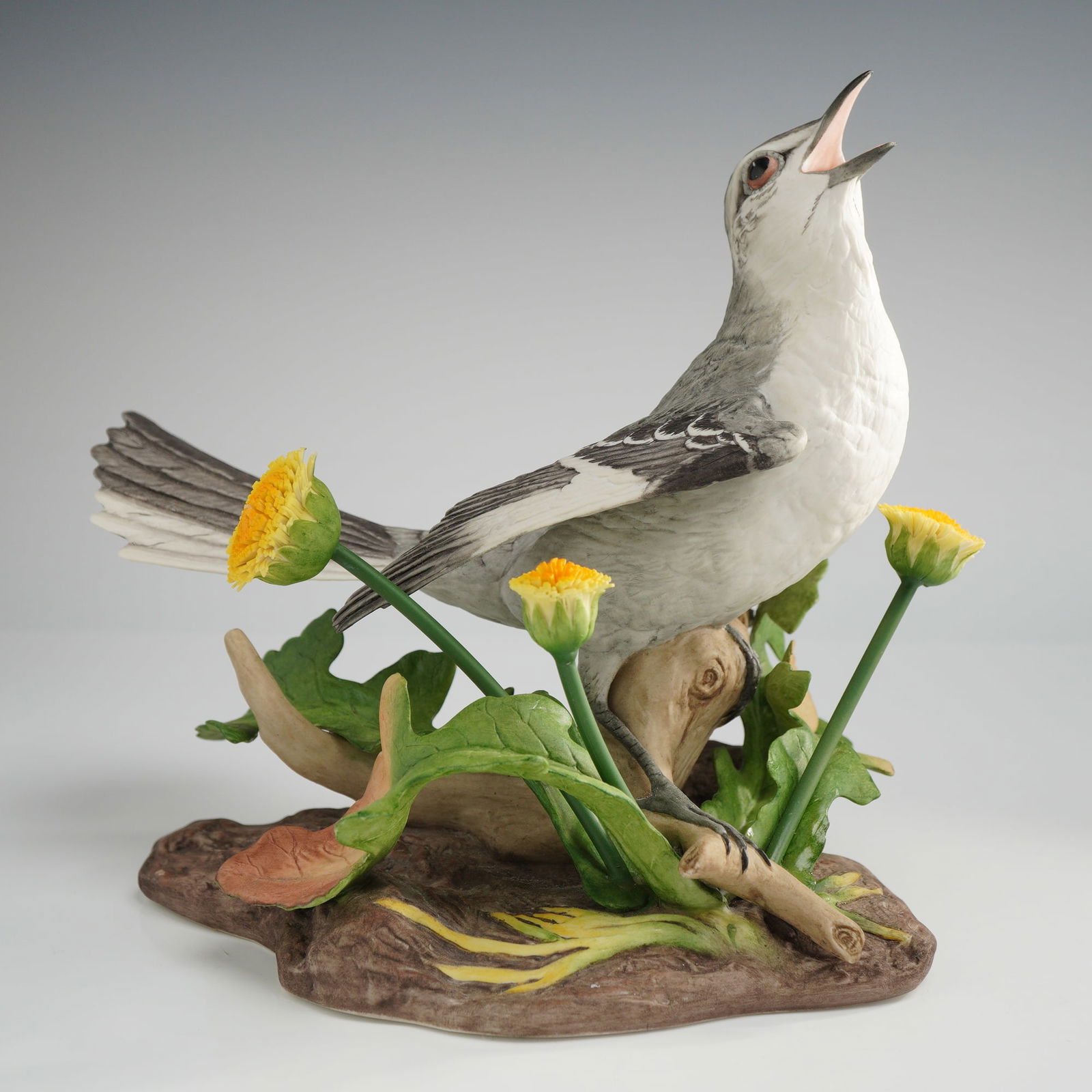 Signed Boehm Porcelain Figurine, Mockingbird 40203: Boehm bisque porcelain figurine of a mockingbird. The hand-painted matte bird is depicted in a singing pose with an open beak, perched on a textured branch and earth-form base. The base is decorated w