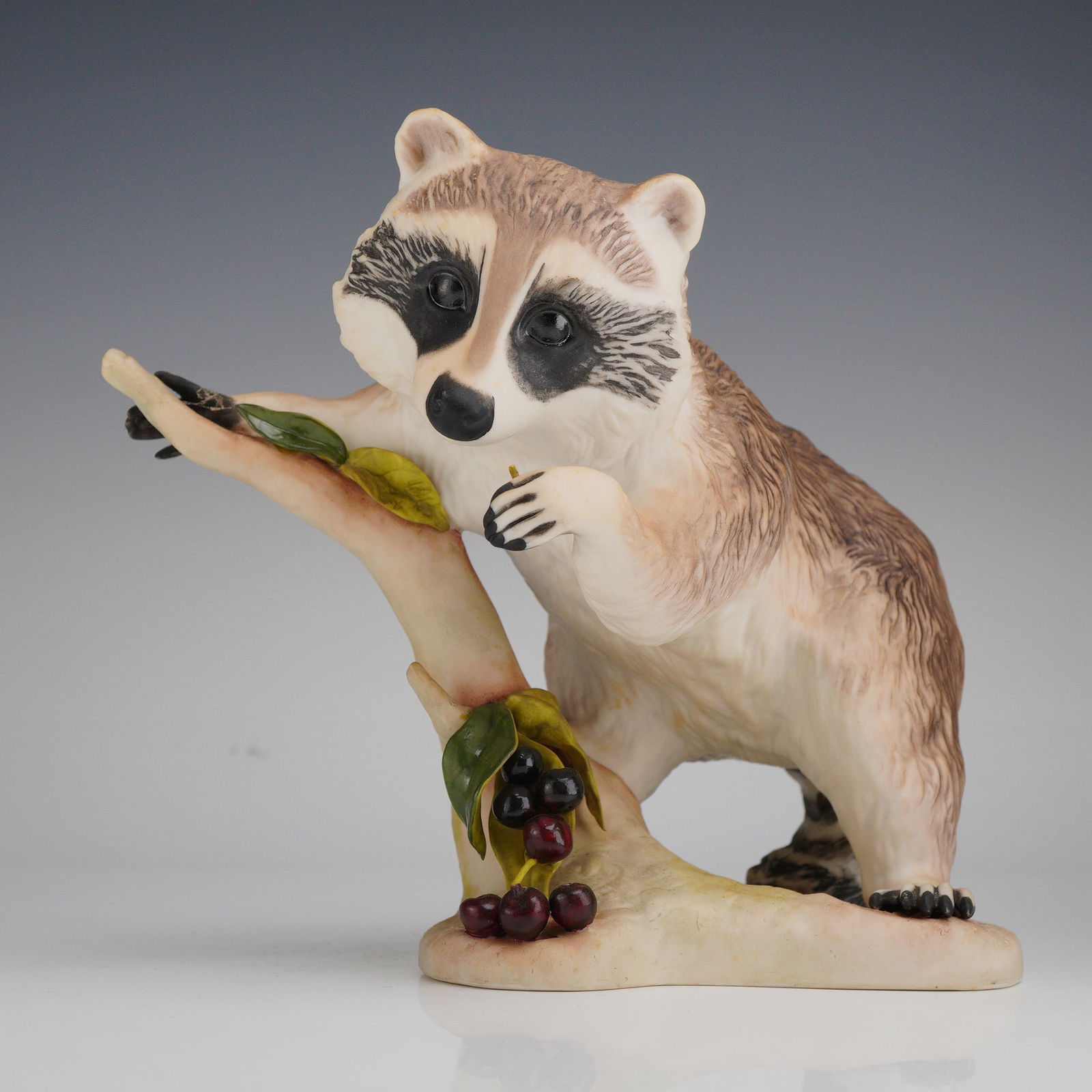 Cybis Porcelain Figurine, Raffles the Raccoon: The sculpture depicts a realistically modeled raccoon climbing a pale branch adorned with green leaves and clusters of dark berries. The piece features a matte bisque finish with detailed textural mod