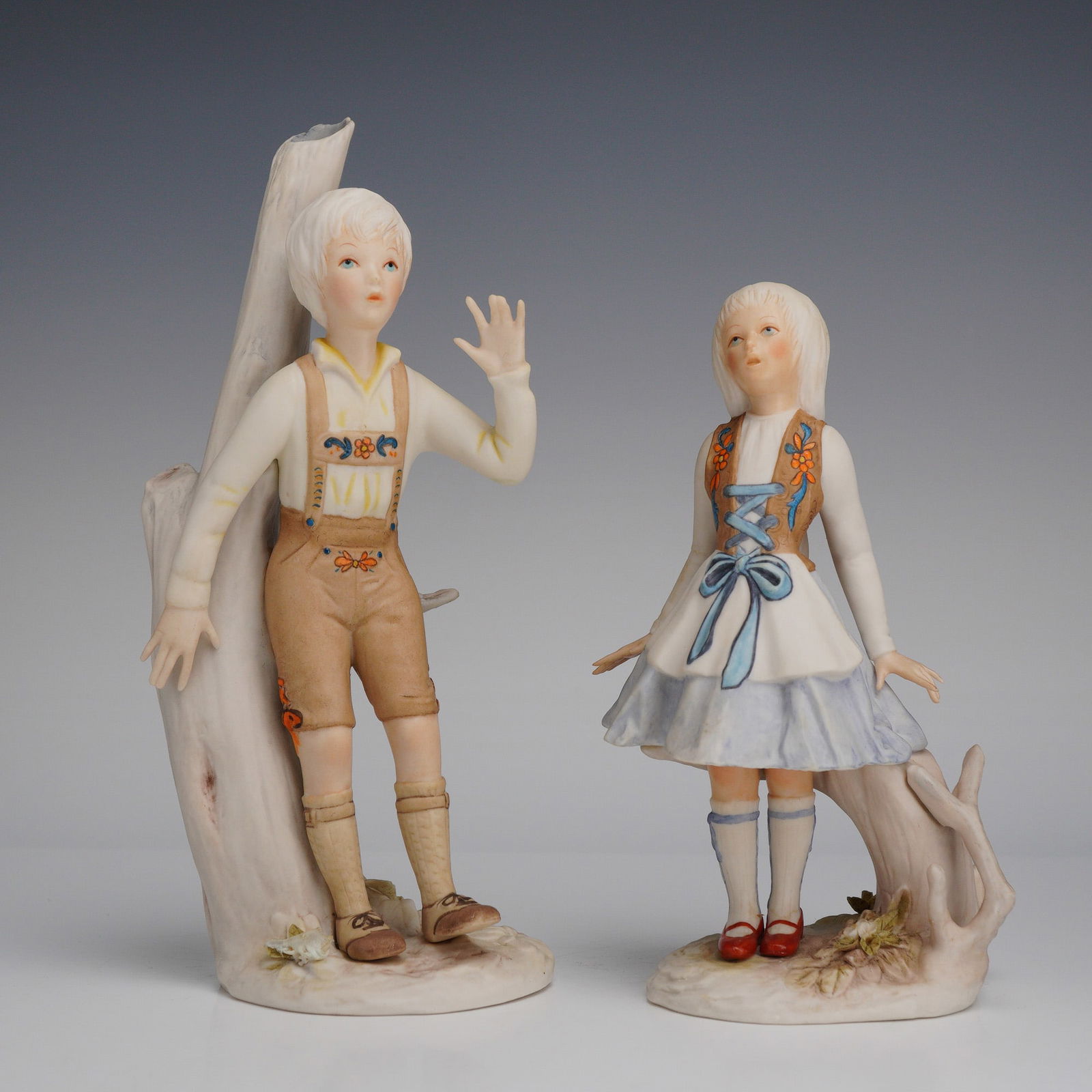 2pc Cybis Bisque Porcelain Figurines, Hansel and Gretel: The Hansel figure is modeled wearing tan lederhosen with floral embroidery, a white shirt, and knee-high socks, leaning against a stylized white tree trunk. The Gretel figure is modeled in a white dre