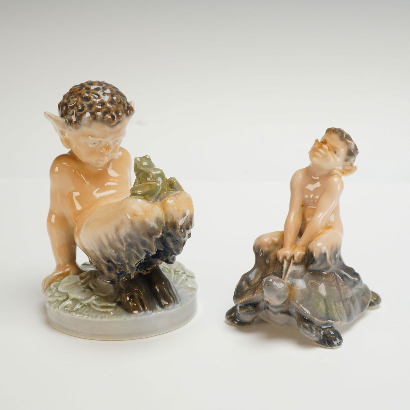 2pc Royal Copenhagen Porcelain Faun Figurines: A pair of Royal Copenhagen glazed porcelain figurines depicting mythological fauns. The larger figurine, marked with model number 1713, portrays a seated faun peering down at a small frog resting on i