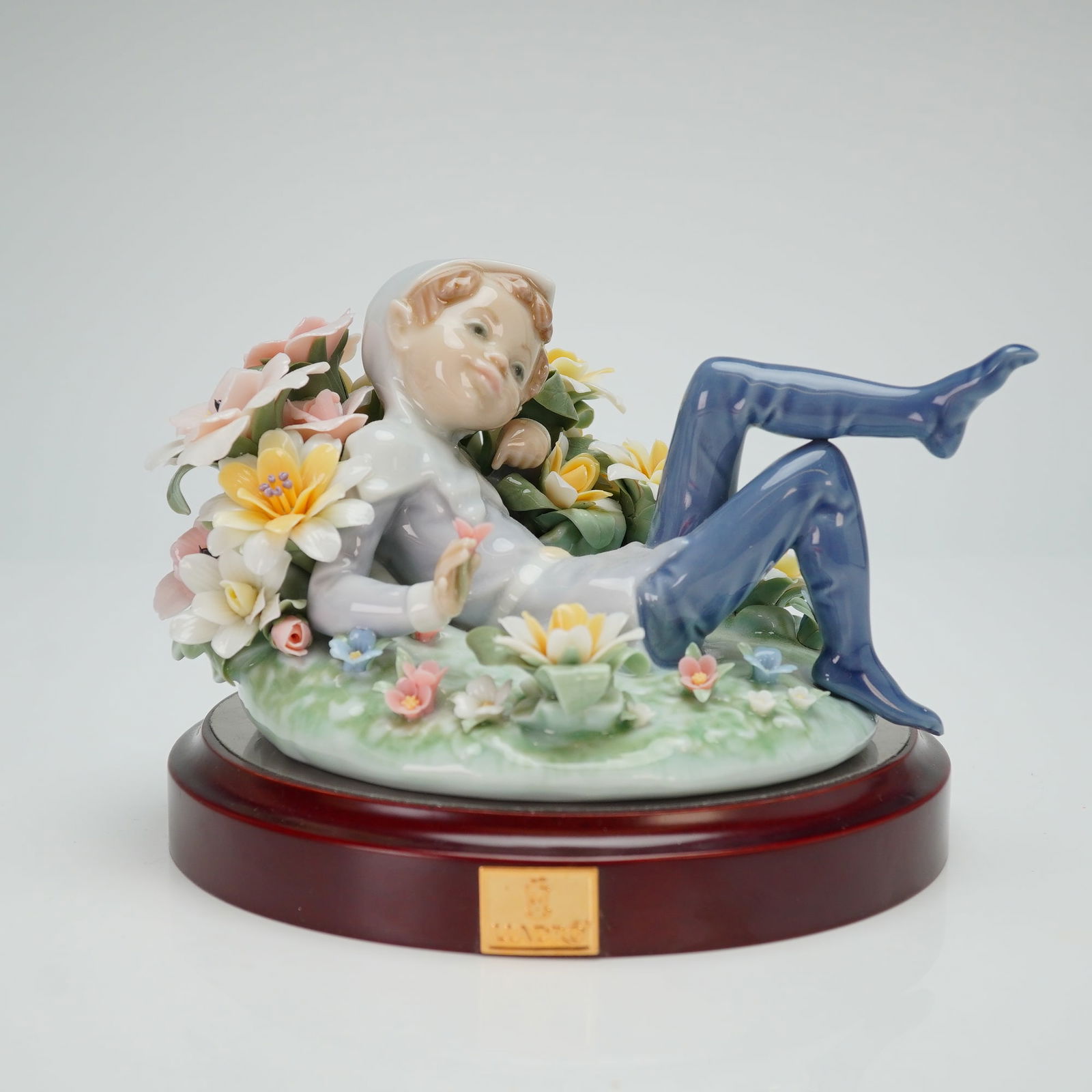 Lladro Porcelain Figurine Leprechaun 01001721 with Base: This handcrafted Spanish porcelain piece depicts a reclining figure in a hooded gray garment and blue leggings, resting among a profusion of delicately modeled flowers in shades of pink, yellow, and w