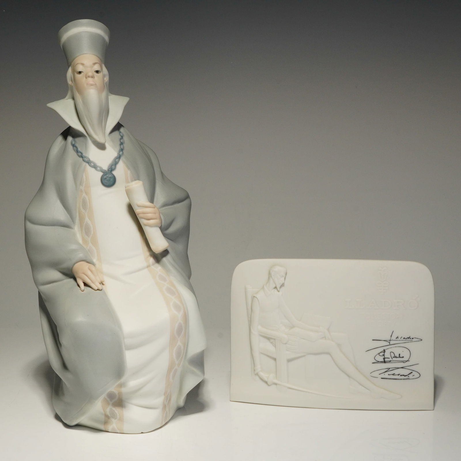 Two Lladro Matte Porcelain Figurine, Lawyer 01011089 and Plaque: A pair of Lladro porcelain items featuring the figurine titled Lawyer or Magistrado, model number 01011089, designed by Juan Huerta. The figurine depicts a seated scholar or legal professional with a