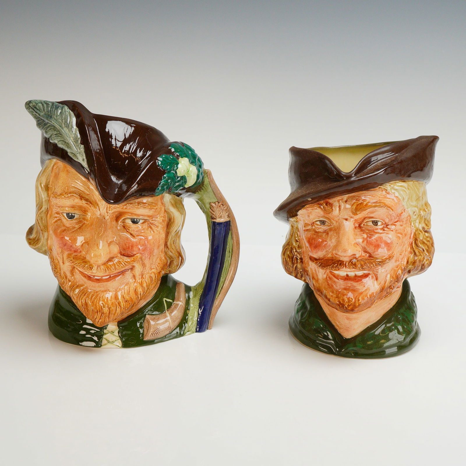 2pc Royal Doulton Character Jugs, Robin Hood D6527 and D6205: Two Royal Doulton glazed ceramic character jugs depicting Robin Hood, consisting of models D6527 and D6205. Each piece features hand-painted details including reddish-blonde beards and traditional gre