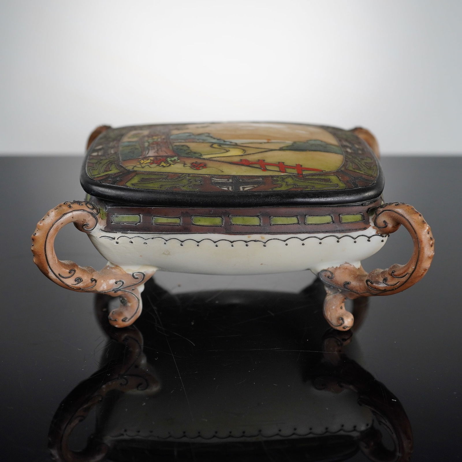 Morimura Bros. Nippon Porcelain Lidded Vanity Box: This hand-painted Morimura Nippon porcelain lidded vanity box features a pastoral landscape scene on the lid depicting a winding path, a red fence, and a flowering tree. The central image is framed
